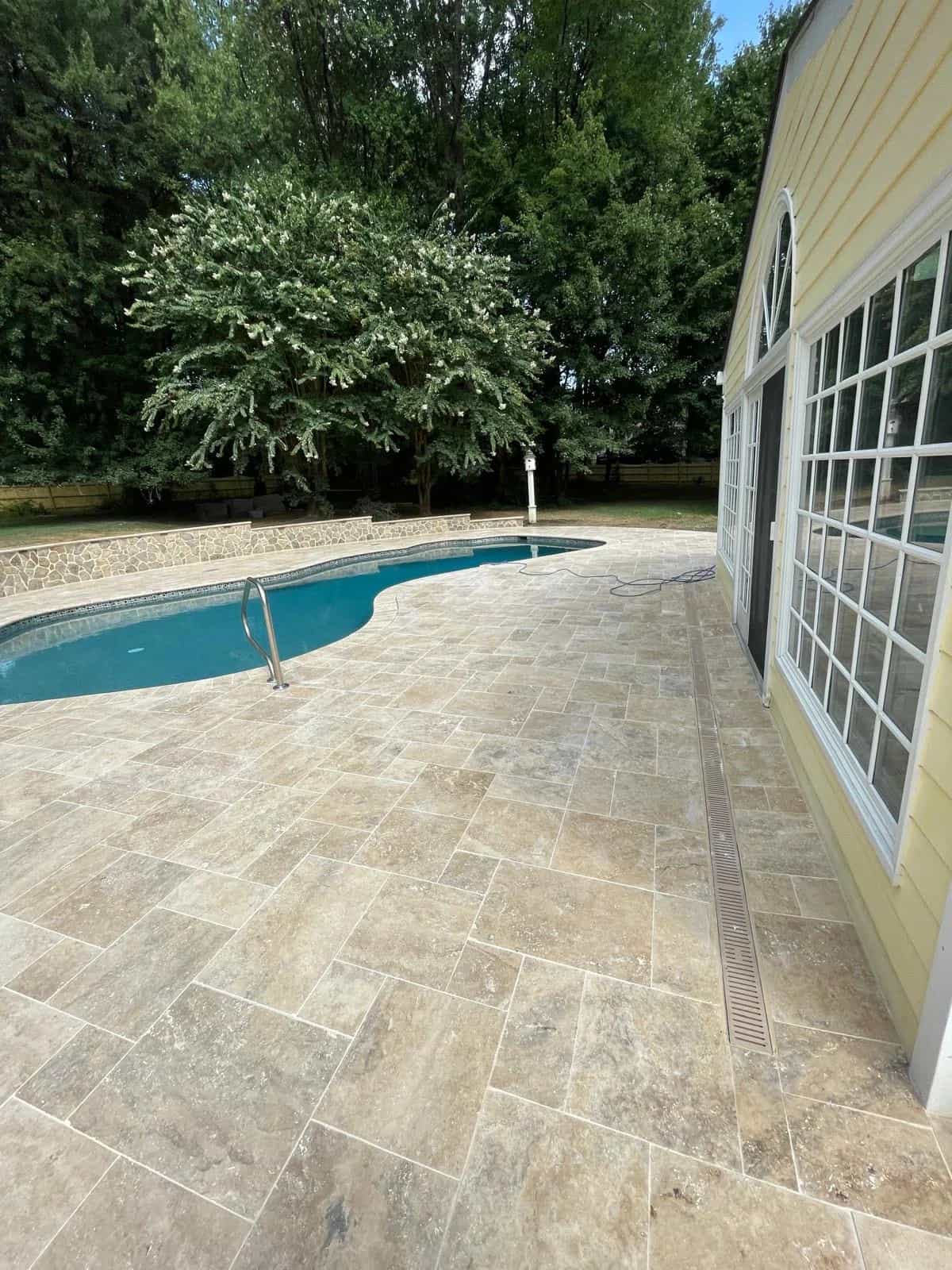 Philly Travertine Tumbled French Pattern 3cm Paver installation showcases a luxurious pool deck. The natural stone's earthy tones complement the blue pool, surrounded by lush greenery and a traditional-style building with large windows, creating a sere...