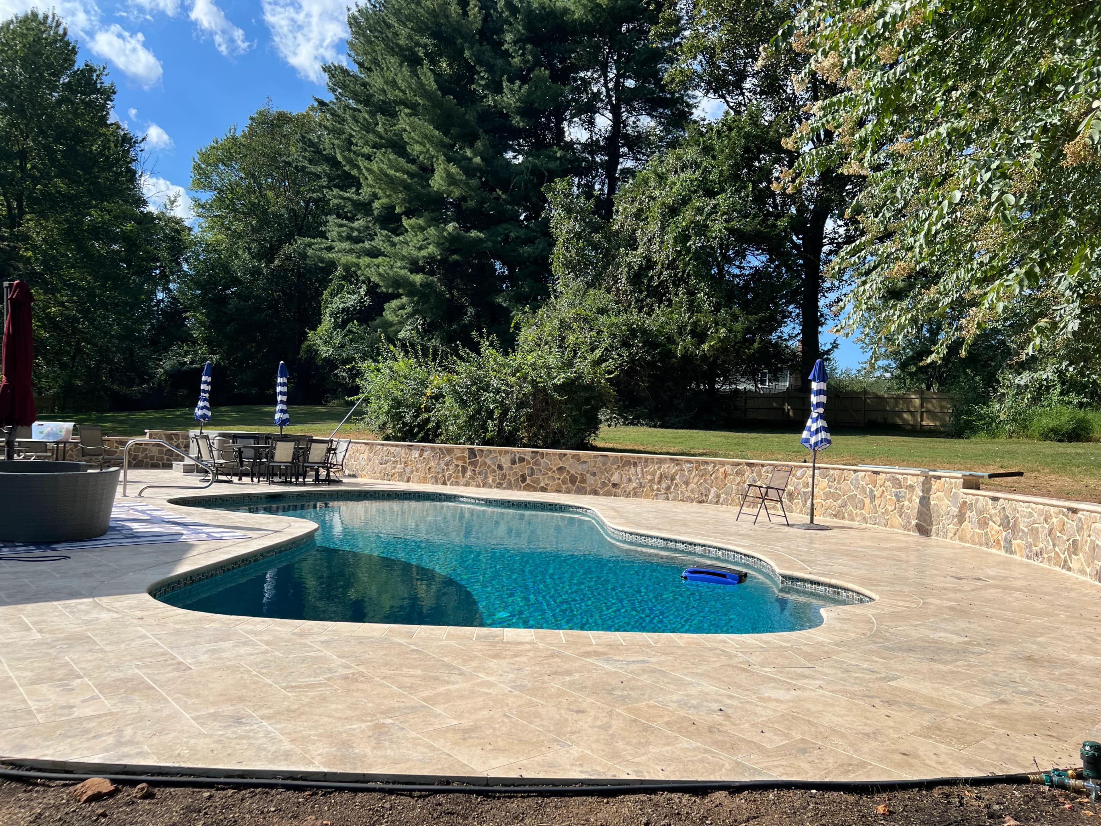 Outdoor pool area featuring Philly Travertine Tumbled French Pattern 3cm Paver - Premium, installed as pool deck. The light beige travertine complements the clear blue pool water and natural stone wall, surrounded by lush green trees and outdoor furnit...