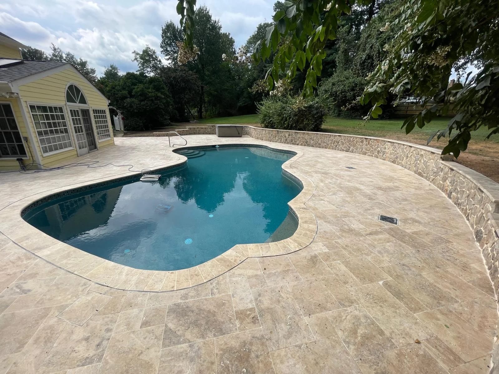 Luxurious installation of Philly Travertine Tumbled French Pattern 3cm Paver surrounds a sparkling pool. The natural stone's creamy tones and textured finish blend with the lush garden and traditional architecture, creating a serene and elegant outdoor...