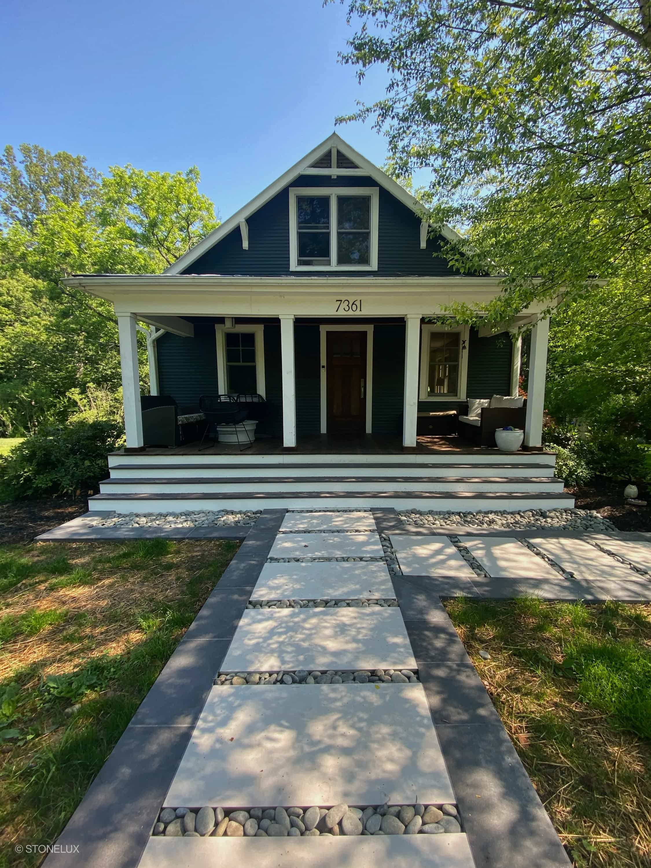 Front entrance showcasing Overland Pearl Matte 24x24x2cm Rectified Porcelain Paver path leading to a traditional dark-painted house. The pavers are interspersed with decorative stones, surrounded by lush landscaping, creating a modern yet classic aesth...