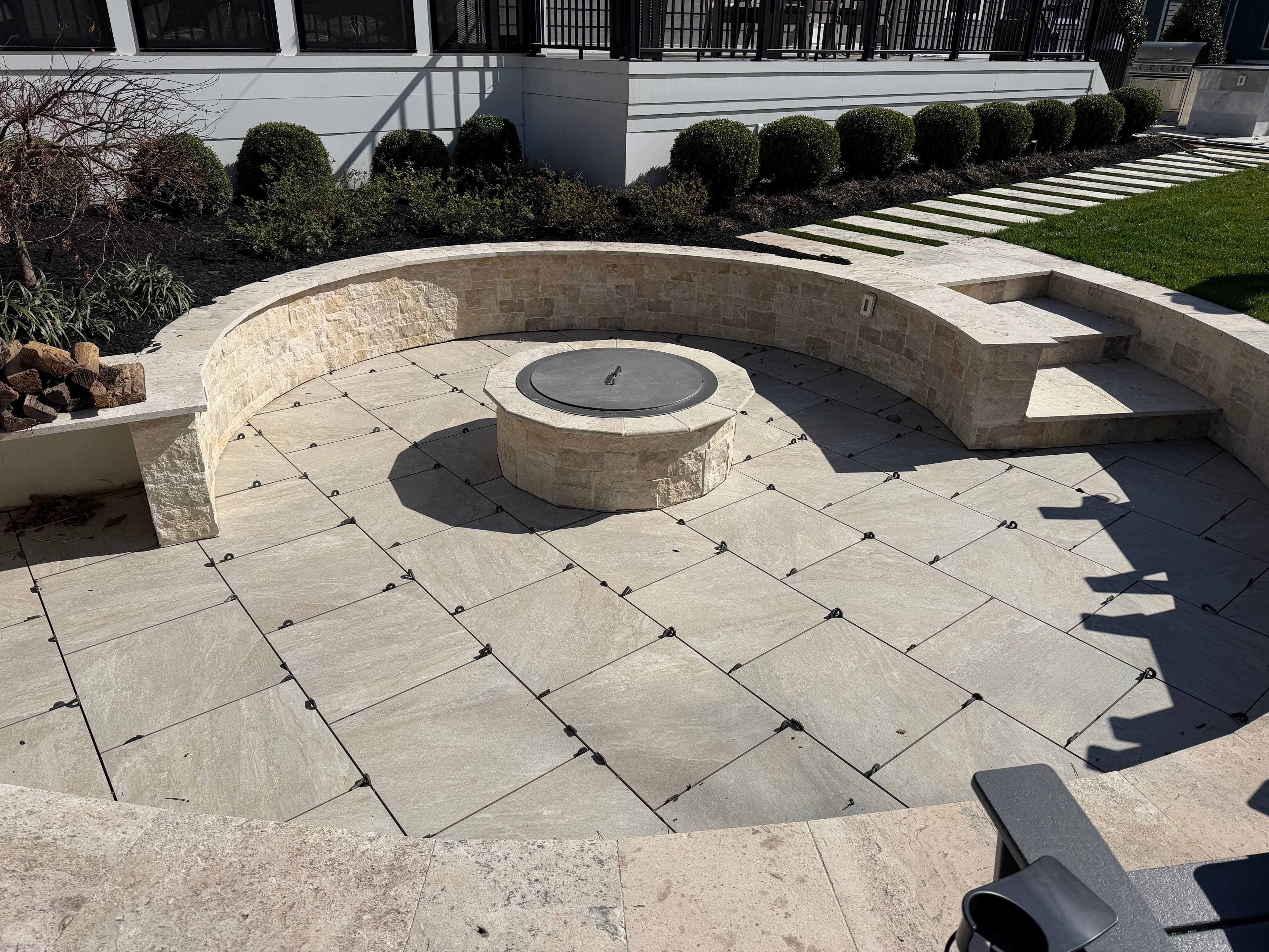 Outdoor patio featuring Rasa Beige Matte 24x24x2cm Rectified Porcelain Paver by STONELUX, laid in a circular pattern around a central stone fire pit. Surrounded by manicured hedges and stone seating, creating a modern, elegant hardscape design.