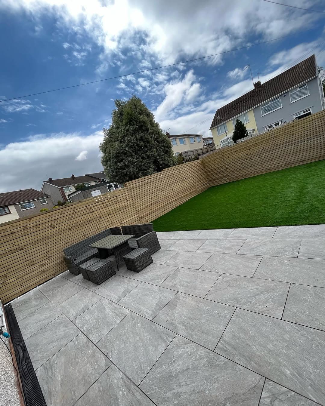 Outdoor patio featuring Rasa Gray Matte 24x36x2cm Rectified Porcelain Pavers by STONELUX. The light gray, matte finish complements a modern garden setting with lush green grass, wooden fencing, and wicker outdoor furniture. Ideal for contemporary hards...