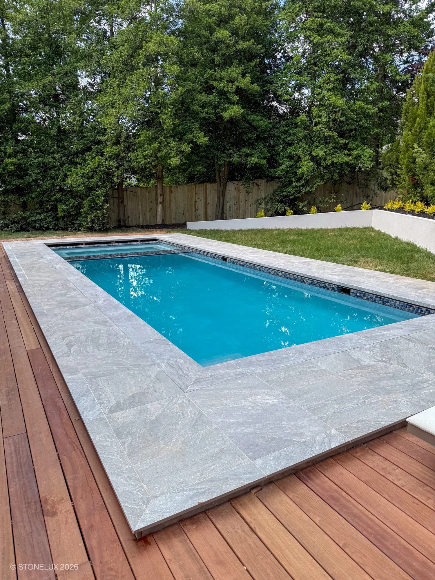 Modern poolside installation featuring Rasa Gray Matte 24x36x2cm Rectified Porcelain Paver as pool coping. The matte finish complements the vibrant blue pool and rich wooden deck. Surrounded by lush greenery, creating a serene outdoor retreat.