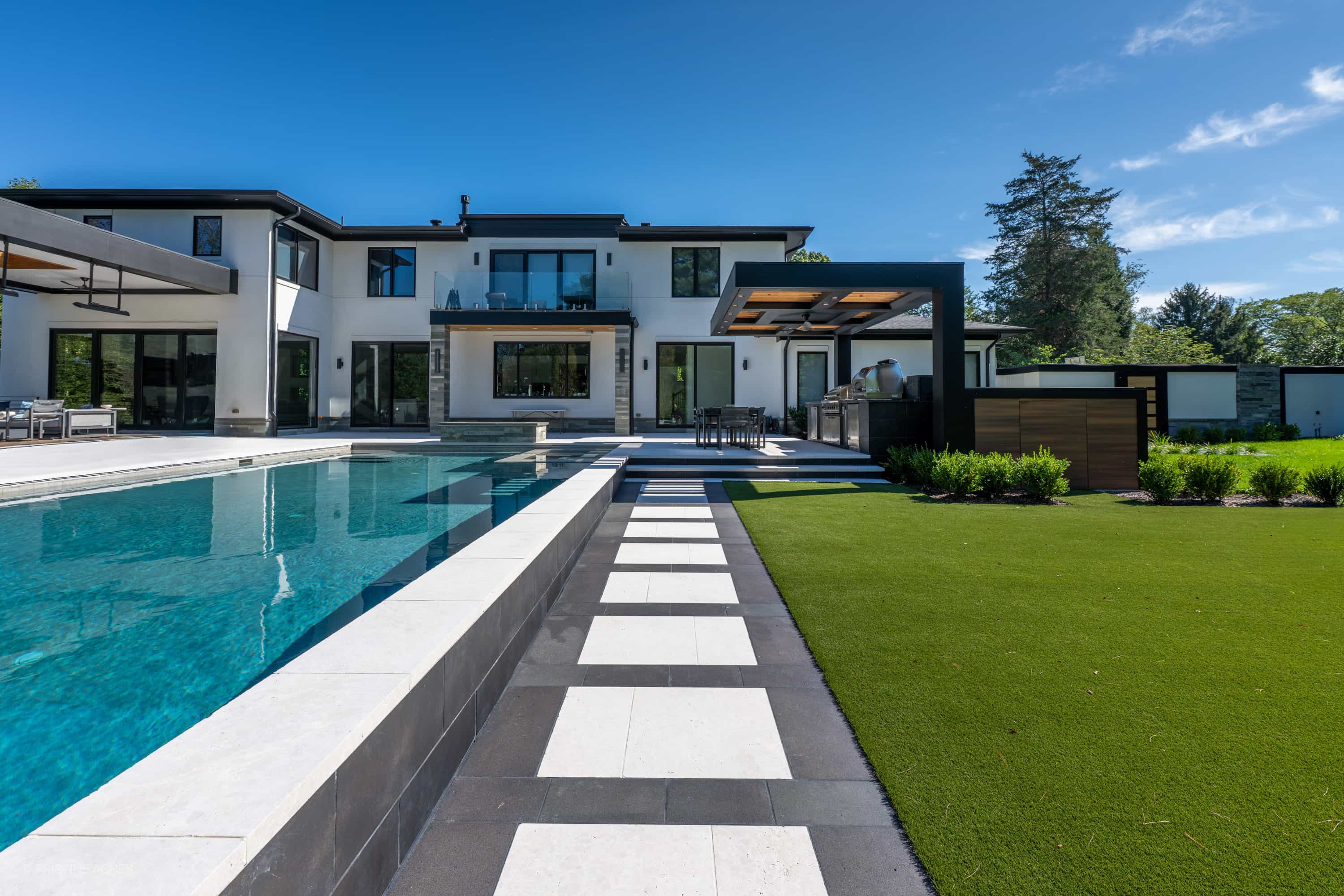 Modern outdoor pool area featuring Black Basalt Tumbled+Brushed 24x24 3cm Pavers by STONELUX. The sleek basalt pavers complement the clear blue pool, contemporary white house, and lush green landscaping, creating a luxurious, cohesive hardscape design.