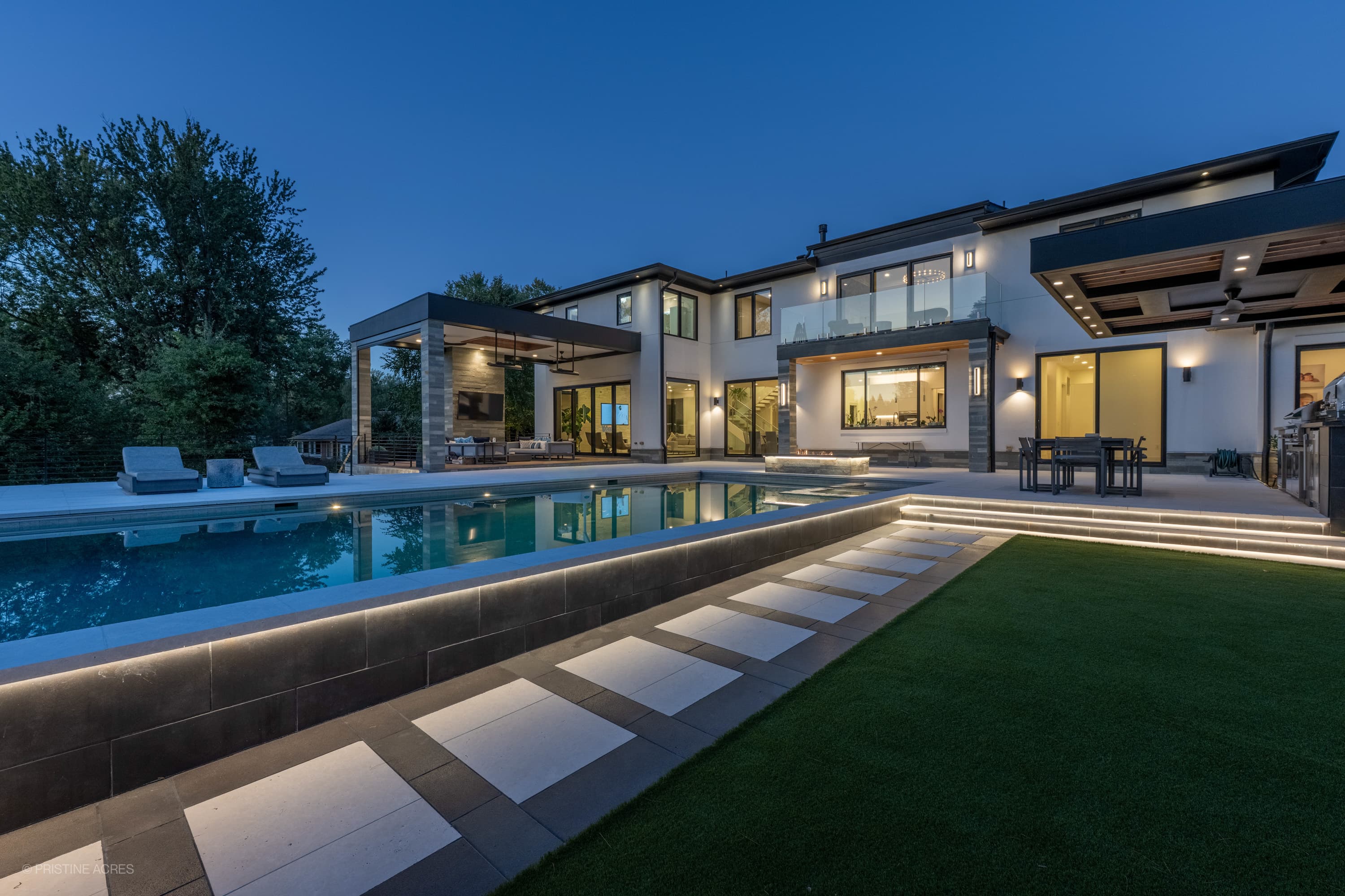 Modern home with Black Basalt Tumbled+Brushed 24x24 3cm Paver patio and pool deck by STONELUX. Features sleek pool, elegant lighting, and stylish outdoor seating, creating a luxurious and contemporary hardscape design surrounded by lush greenery.