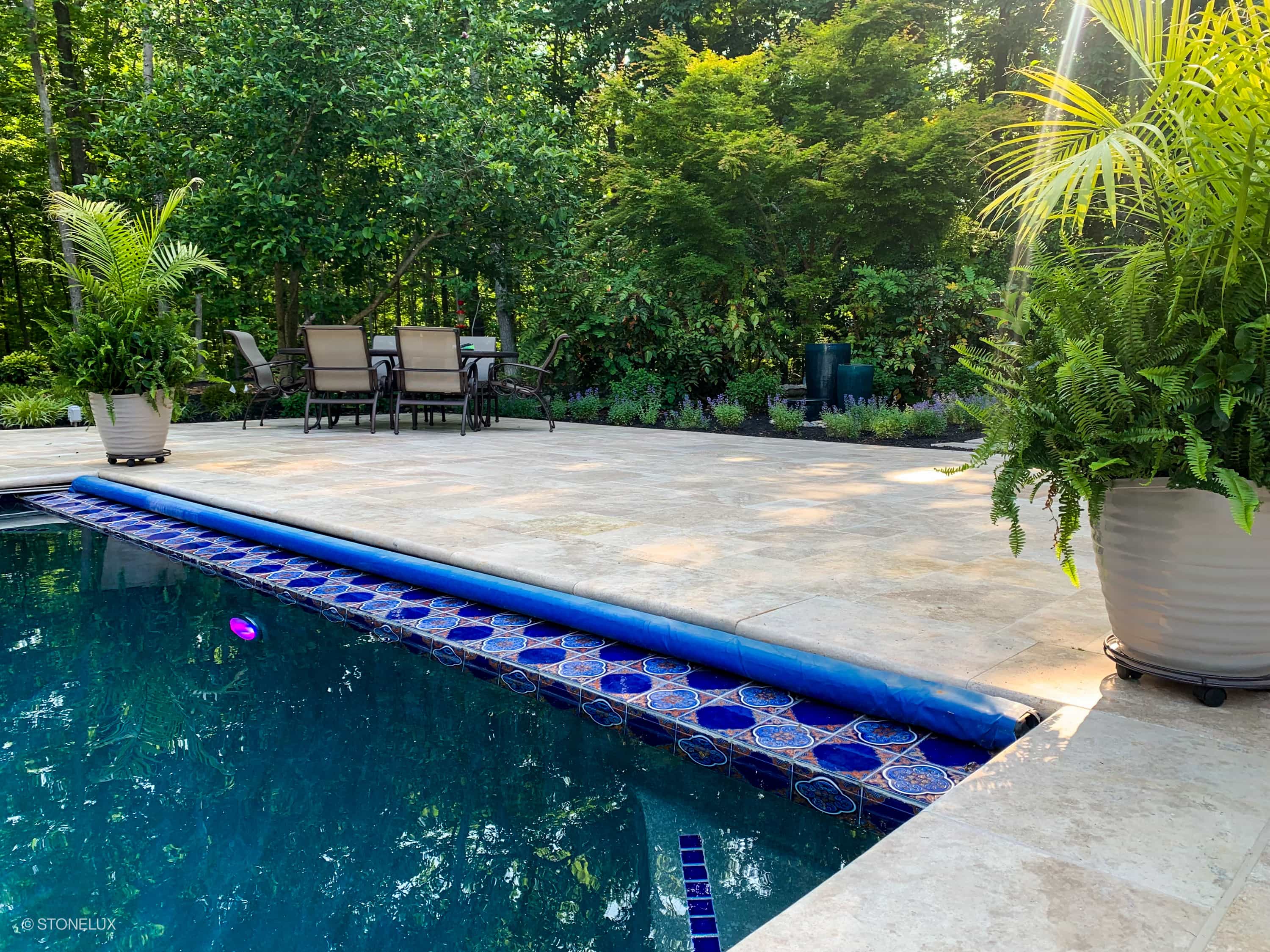 Ivory Travertine Tumbled 14x24x2 Single Bullnose Coping installed around a luxurious pool, featuring light beige travertine with a tumbled finish. Surrounded by lush greenery and elegant patio furniture, creating a serene outdoor retreat. Perfect for p...