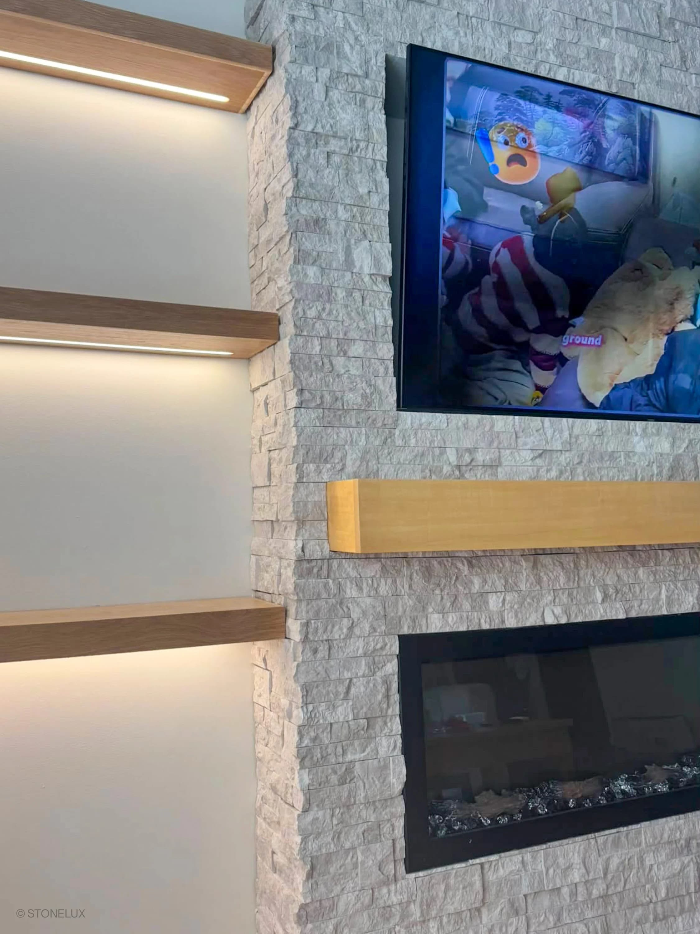 Pera Cream Marble Splitface Ledger Panel - 6x24 installed as a modern fireplace surround. The textured marble veneer complements wooden shelves and a sleek TV setup, creating a contemporary living space with warm, neutral tones and elegant design eleme...