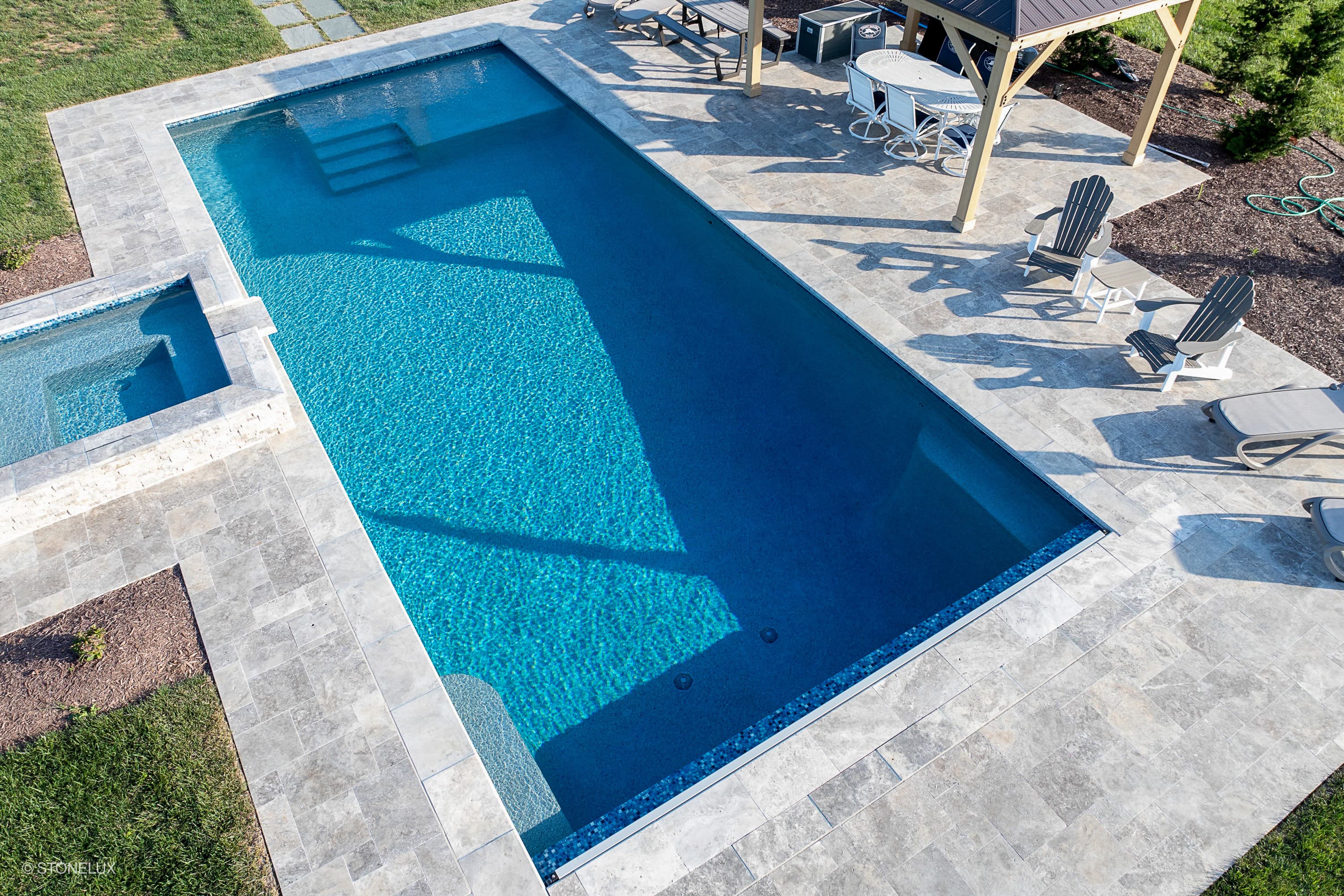 Outdoor pool area showcasing Silver Travertine Tumbled French Pattern 3cm Paver by STONELUX. The elegant travertine pavers frame the turquoise pool, complementing modern patio furniture under a pergola. Ideal for luxurious pool deck designs.