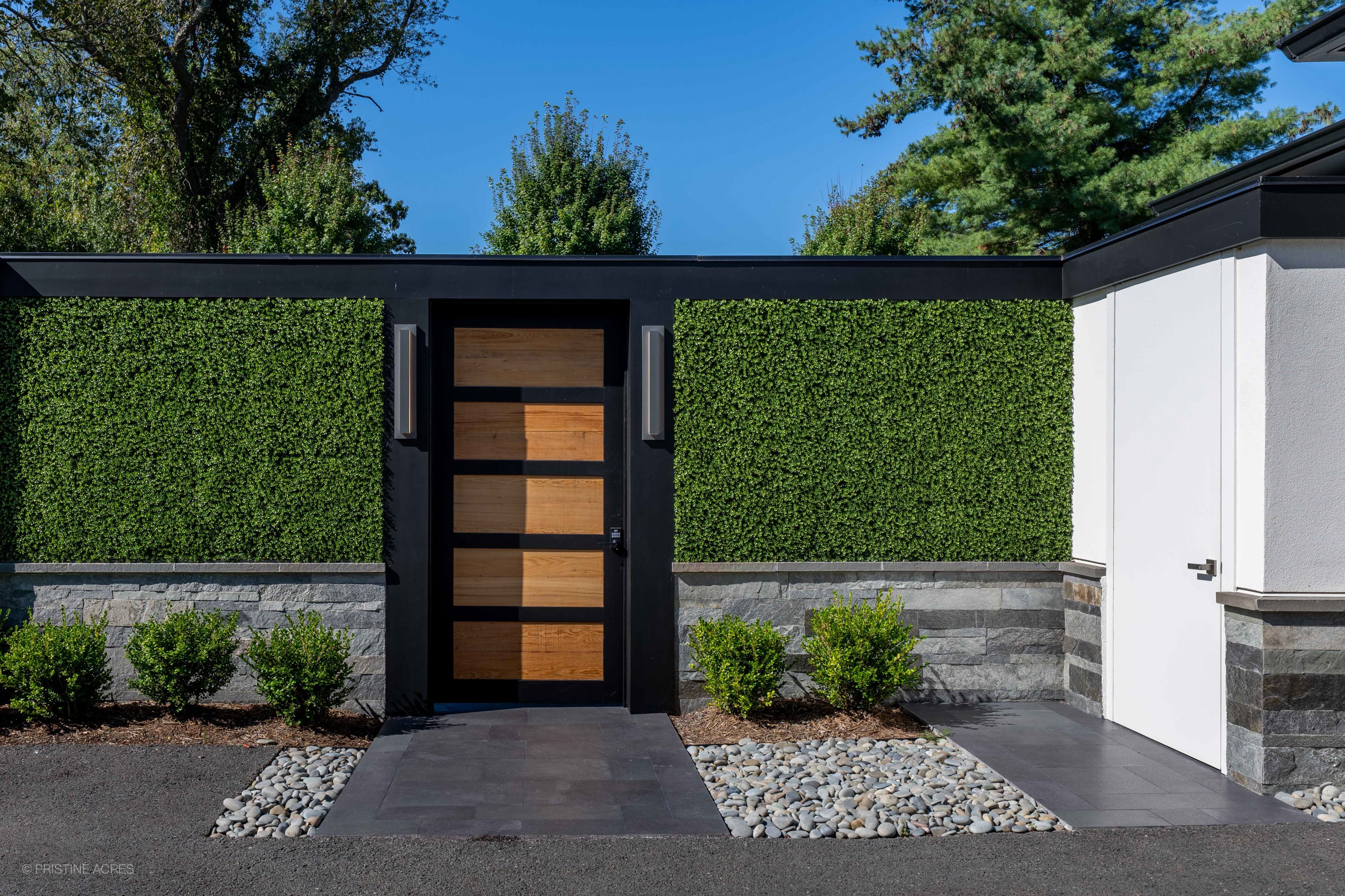 Modern exterior with Black Basalt Tumbled+Brushed 24x24 3cm Paver walkway by STONELUX. The sleek basalt pavers enhance the contemporary design featuring lush greenery, stone walls, and a wooden accent door, creating an elegant, cohesive outdoor aesthetic.