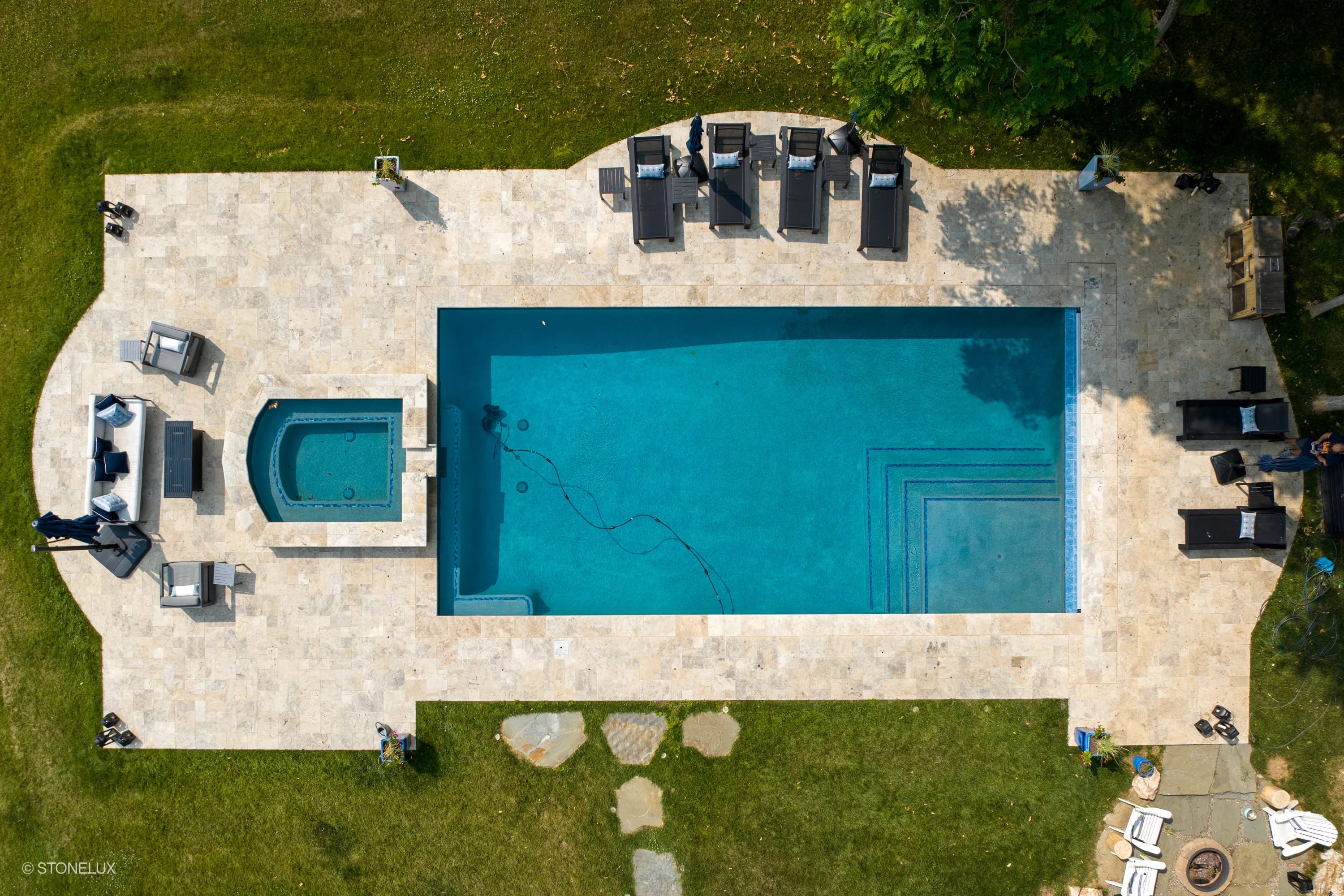 Aerial view of a luxurious pool deck featuring Philly Travertine Tumbled French Pattern 3cm Paver - Premium by STONELUX. The travertine's natural beige tones complement the vibrant blue pool and lush green surroundings. Elegant lounge chairs and a hot ...