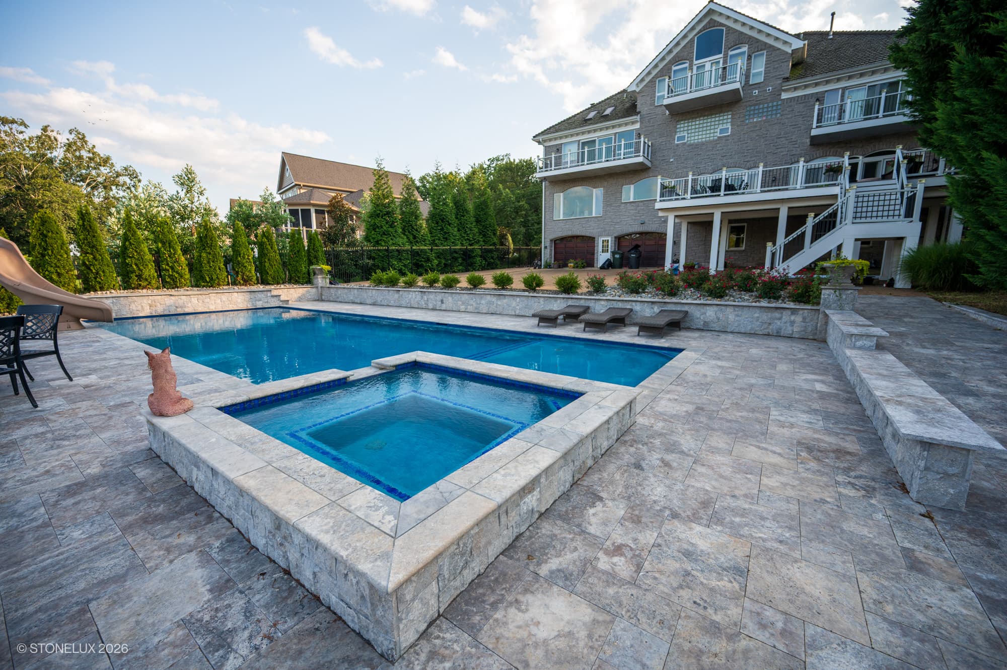 Luxury pool area featuring Silver Titanium Travertine Tumbled French Pattern 3cm Paver by STONELUX. The travertine's natural texture and gray tones enhance the modern pool deck, complemented by lush greenery and elegant outdoor furniture, perfect for p...