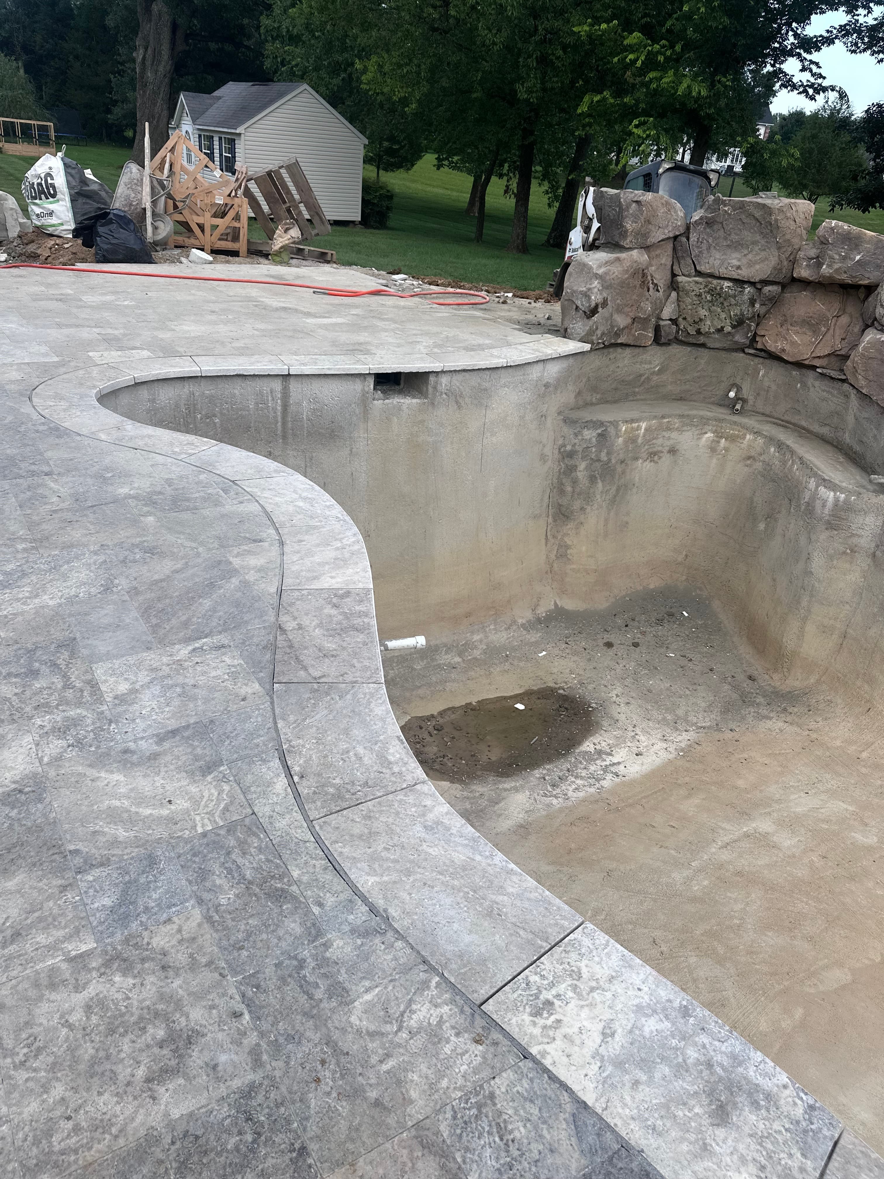 Silver Travertine Tumbled French Pattern 3cm Paver installed as pool deck in a hardscape project. The natural stone's subtle gray tones and tumbled finish complement the rustic stone wall and unfinished pool, set in a lush green landscape.