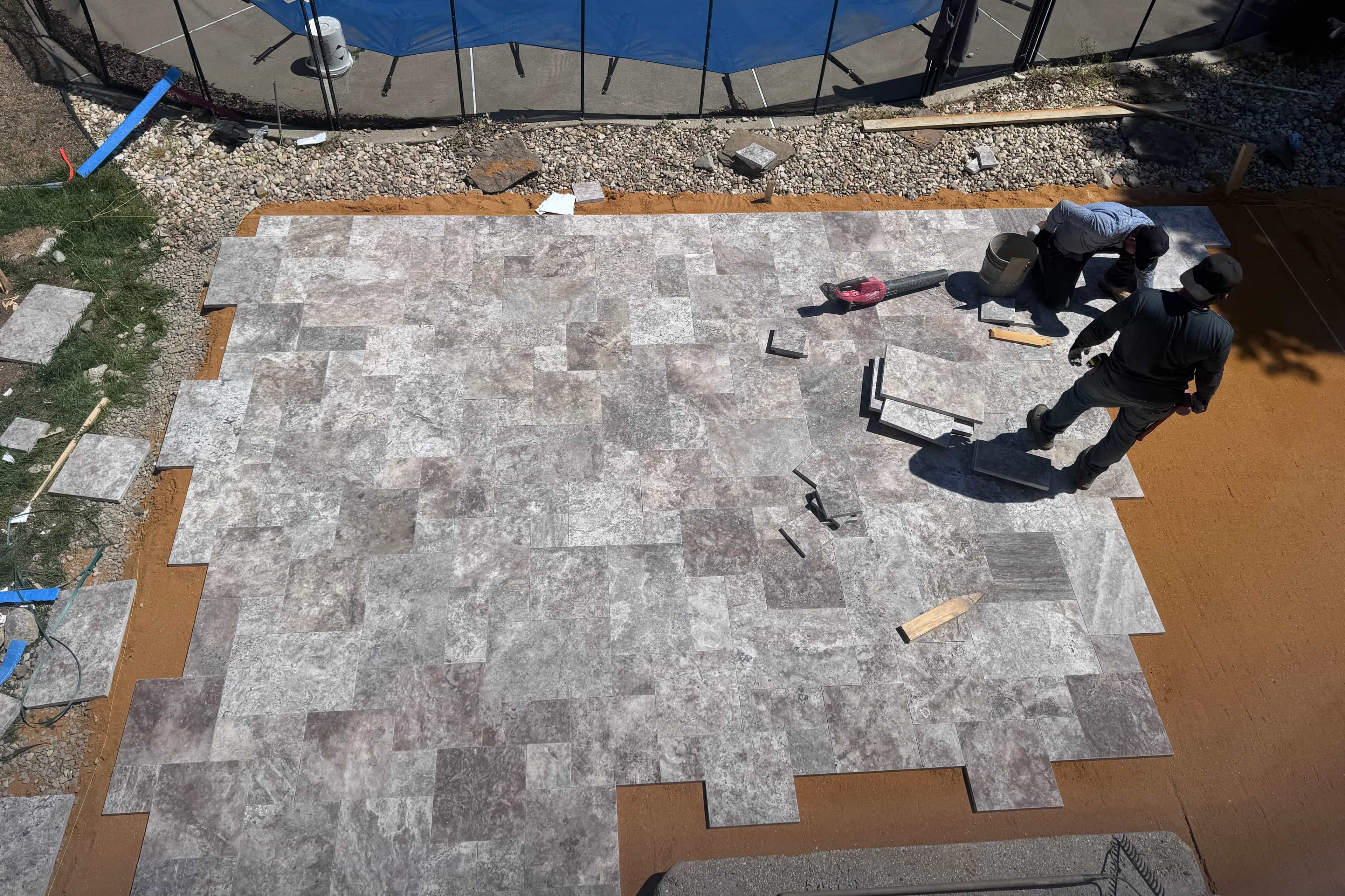 Installation of Silver Titanium Travertine Tumbled French Pattern 3cm Pavers by STONELUX. Workers lay natural stone pavers around a pool area, bordered with gravel and grass. The tumbled finish and varied gray tones create a sophisticated, durable outd...