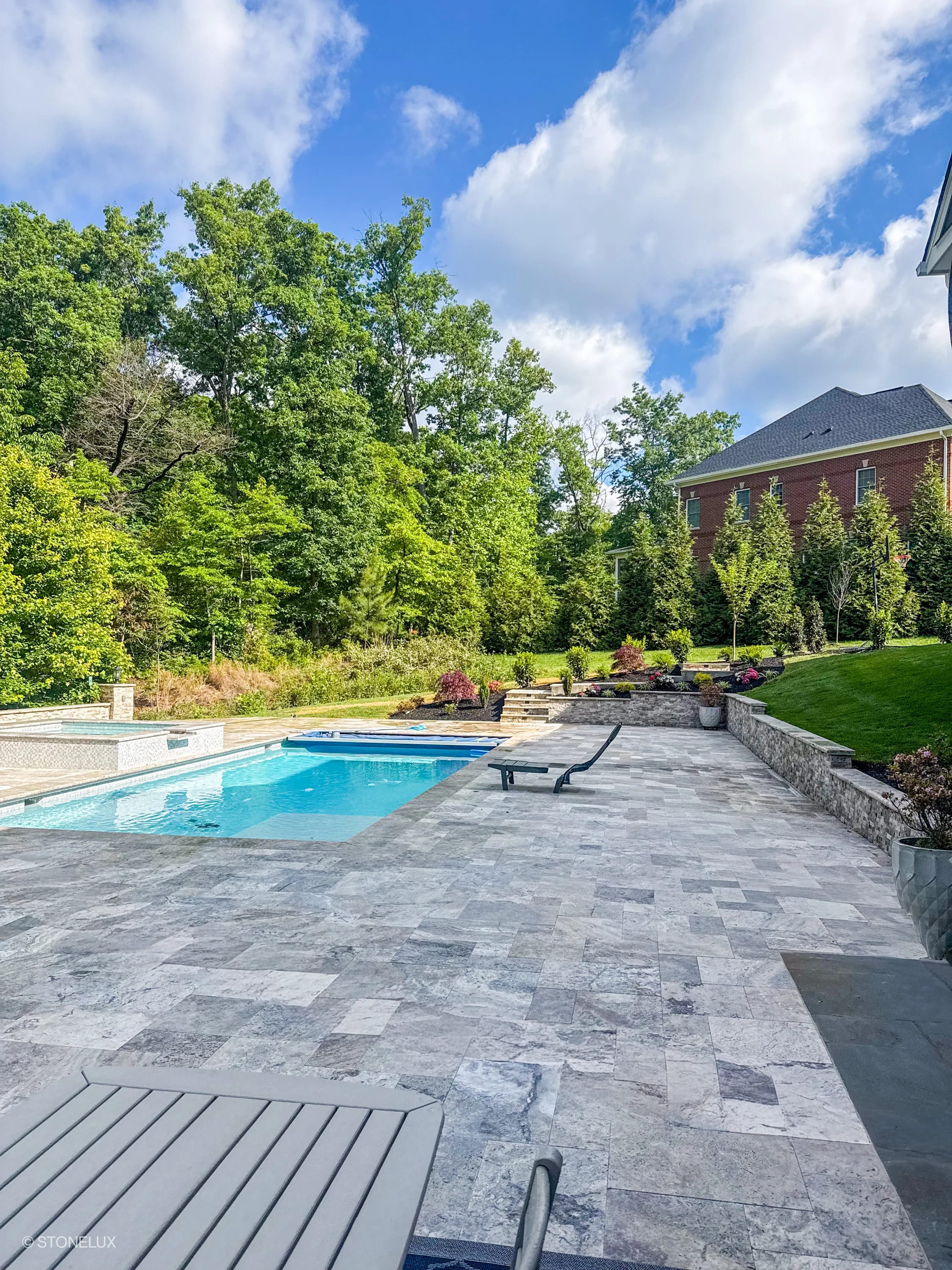 Outdoor pool area featuring Silver Travertine Tumbled French Pattern 3cm Paver - Select by STONELUX. The travertine pavers create a stylish patio around a blue pool, surrounded by lush greenery and elegant landscaping, ideal for modern hardscape projects.
