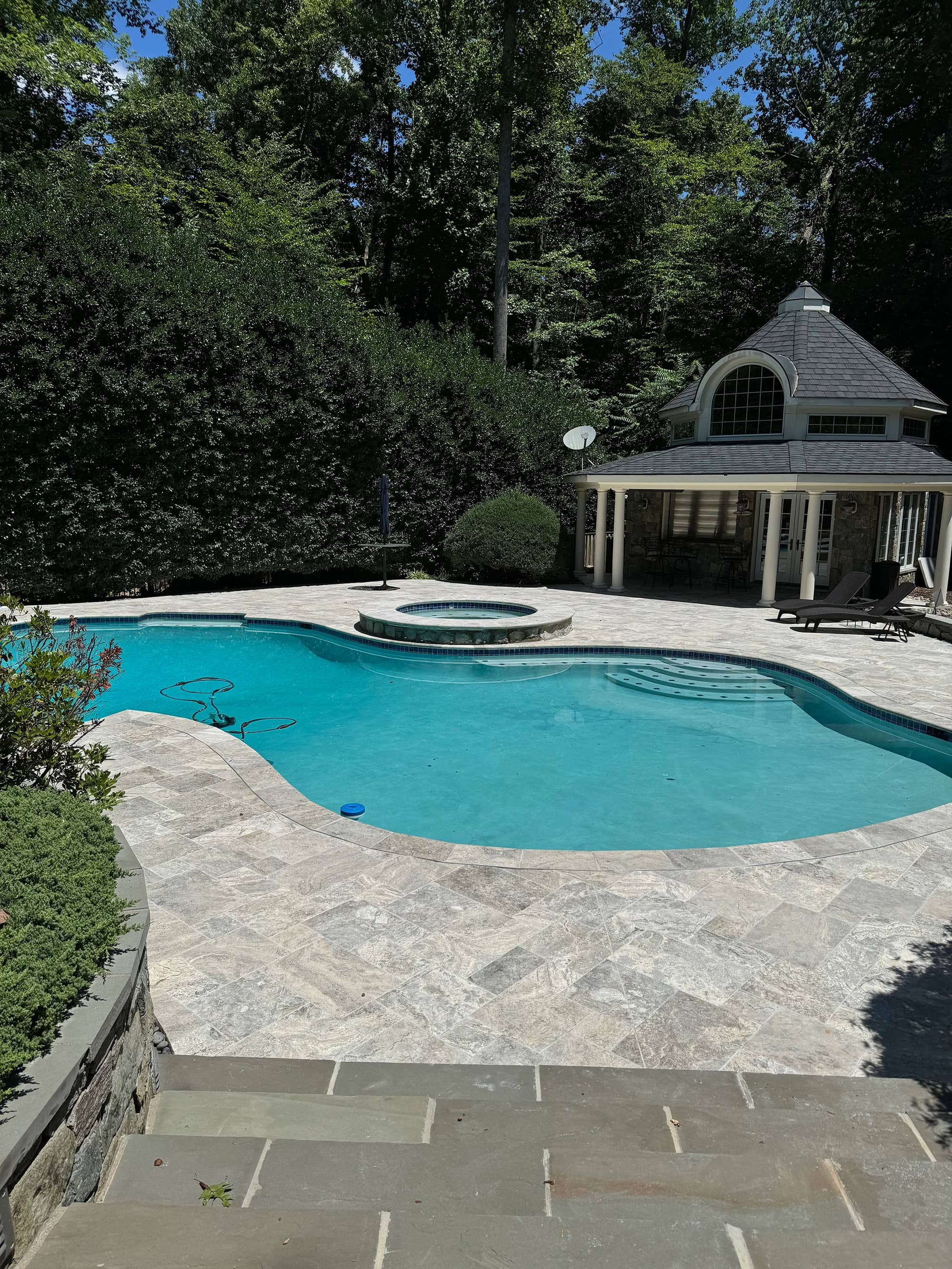 Luxurious poolside featuring Silver Travertine Tumbled French Pattern 3cm Paver by STONELUX. The natural stone pavers create a sophisticated patio with their smooth, tumbled finish. Surrounded by lush greenery, a classic gazebo, and sparkling blue pool...