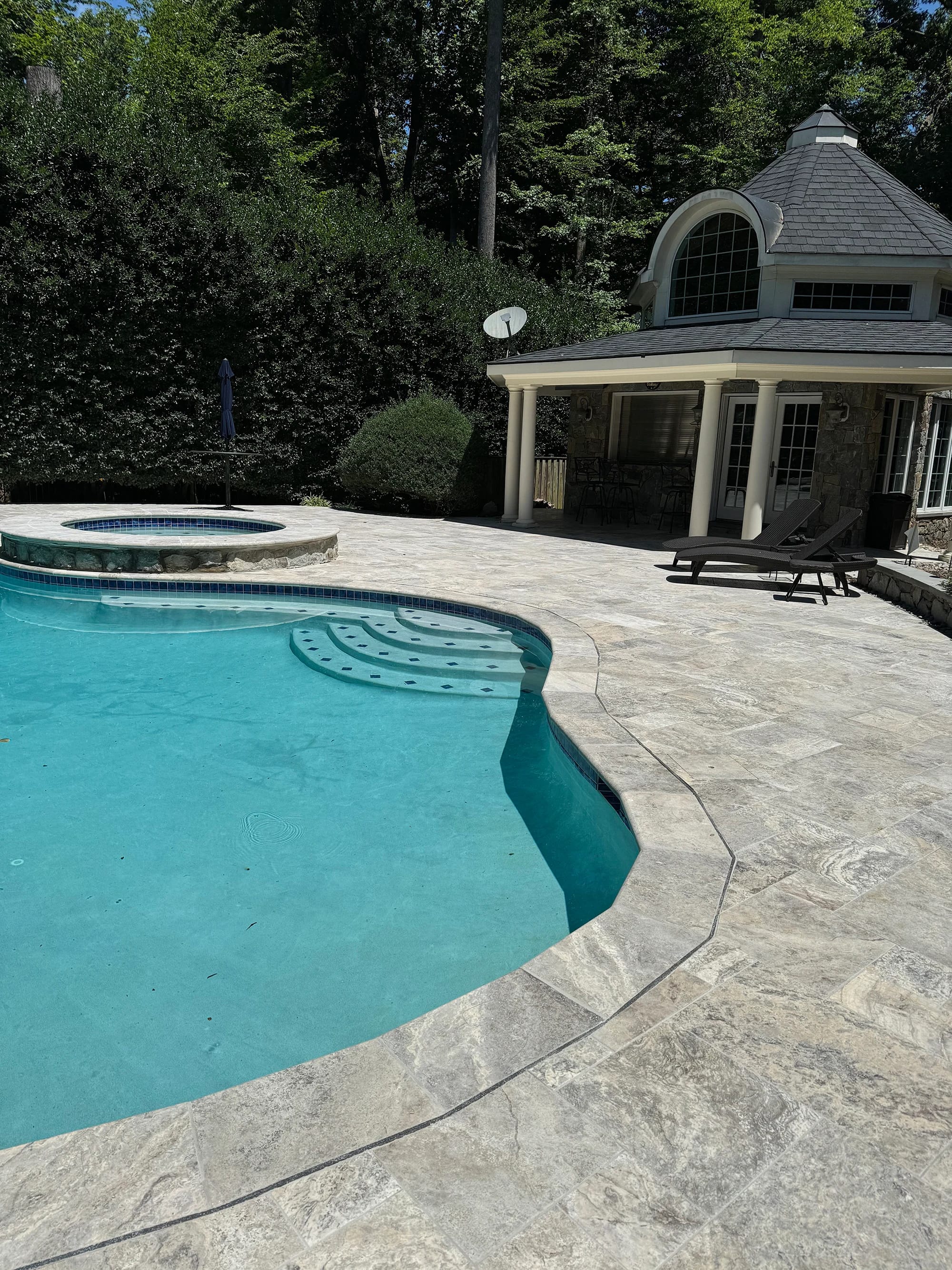 Elegant poolside installation featuring the Silver Travertine Tumbled French Pattern 3cm Paver by STONELUX. The natural stone's subtle gray hues and tumbled finish complement the clear blue pool, lush greenery, and classic architecture, creating a sere...