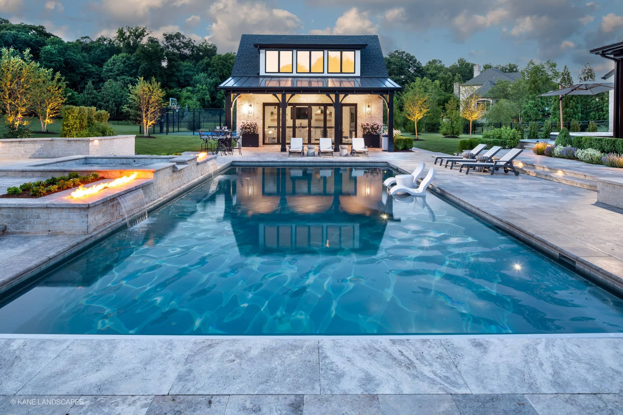 Modern Oasis at Grenata Preserve in Leesburg, VA features a luxurious pool with Silver Travertine Premium pavers. The modern design includes a sleek fire feature, water cascades, and white lounge chairs. Surrounded by lush landscaping and elegant outdo...