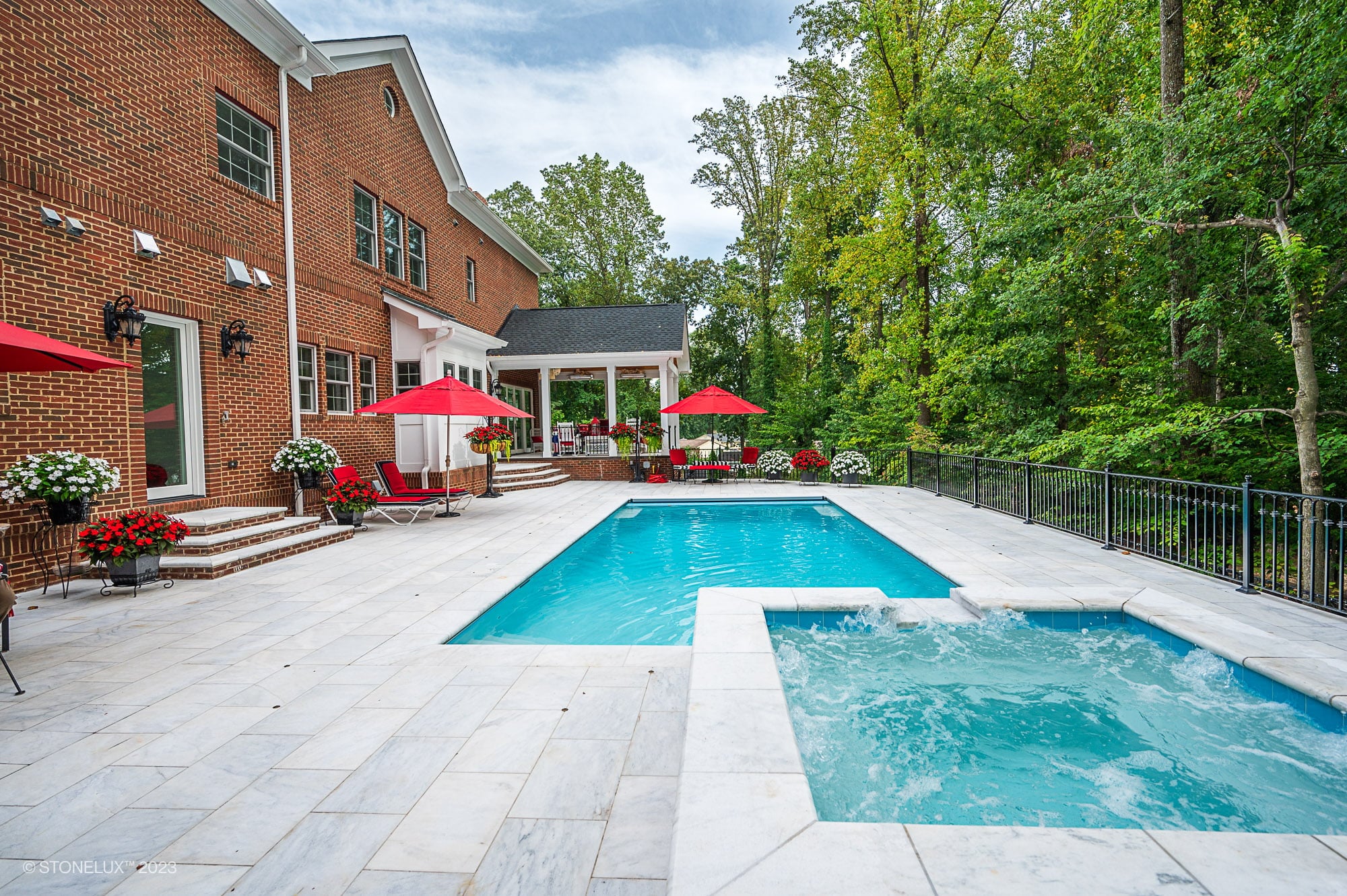 Backyard view of a brick house with a large patio, swimming pool featuring White Whisper: The Pristine Afyon Marble Pool Elegance, hot tub, red umbrellas, lounge chairs, and flower pots, surrounded by trees and a black metal fence.