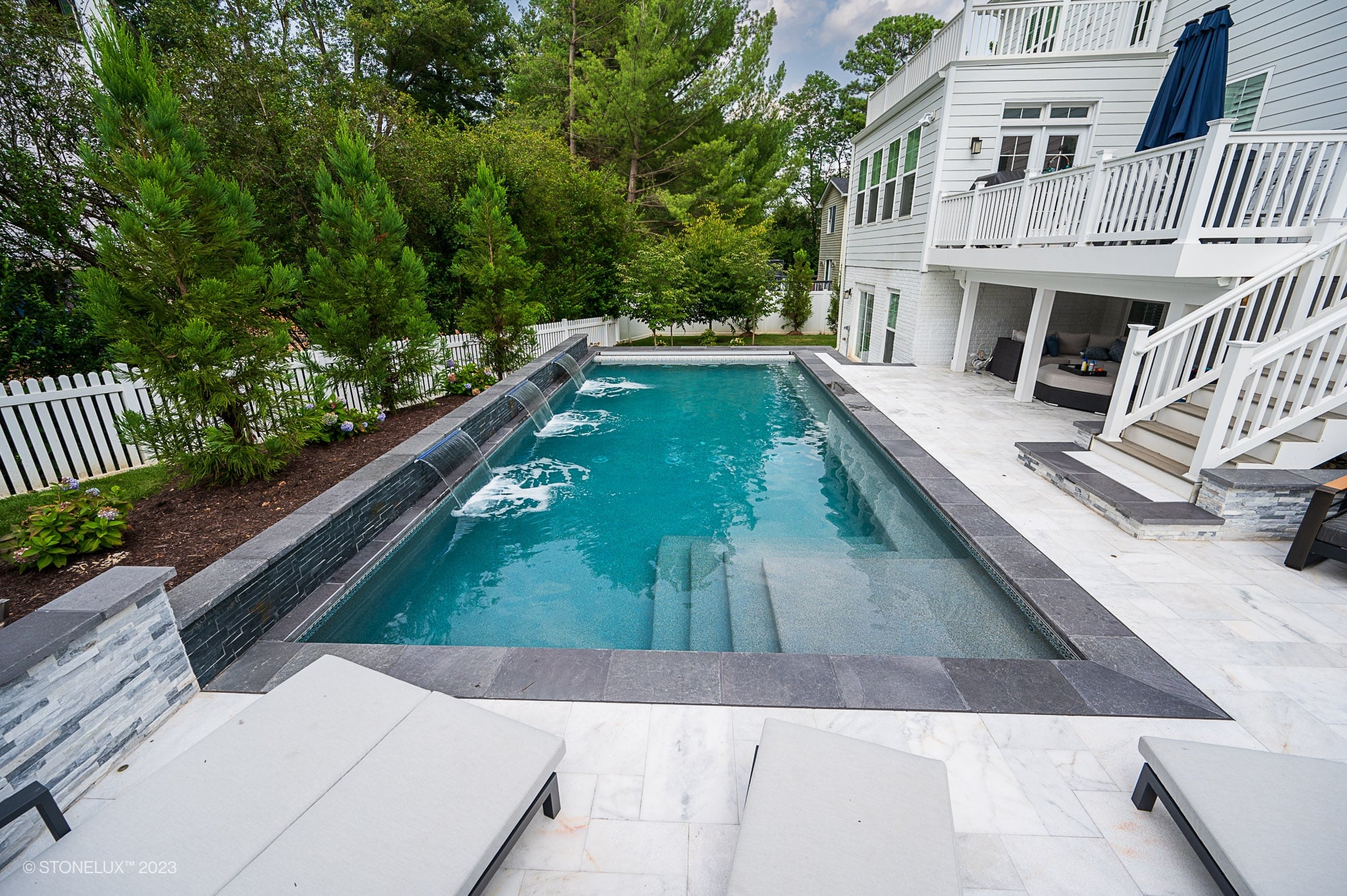 A modern backyard features a rectangular inground pool with built-in steps and water jets, surrounded by Modern Monochrome: The Afyon White Marble & Black Basalt Poolside Harmony, lounge chairs, and a white house with a balcony and covered patio.