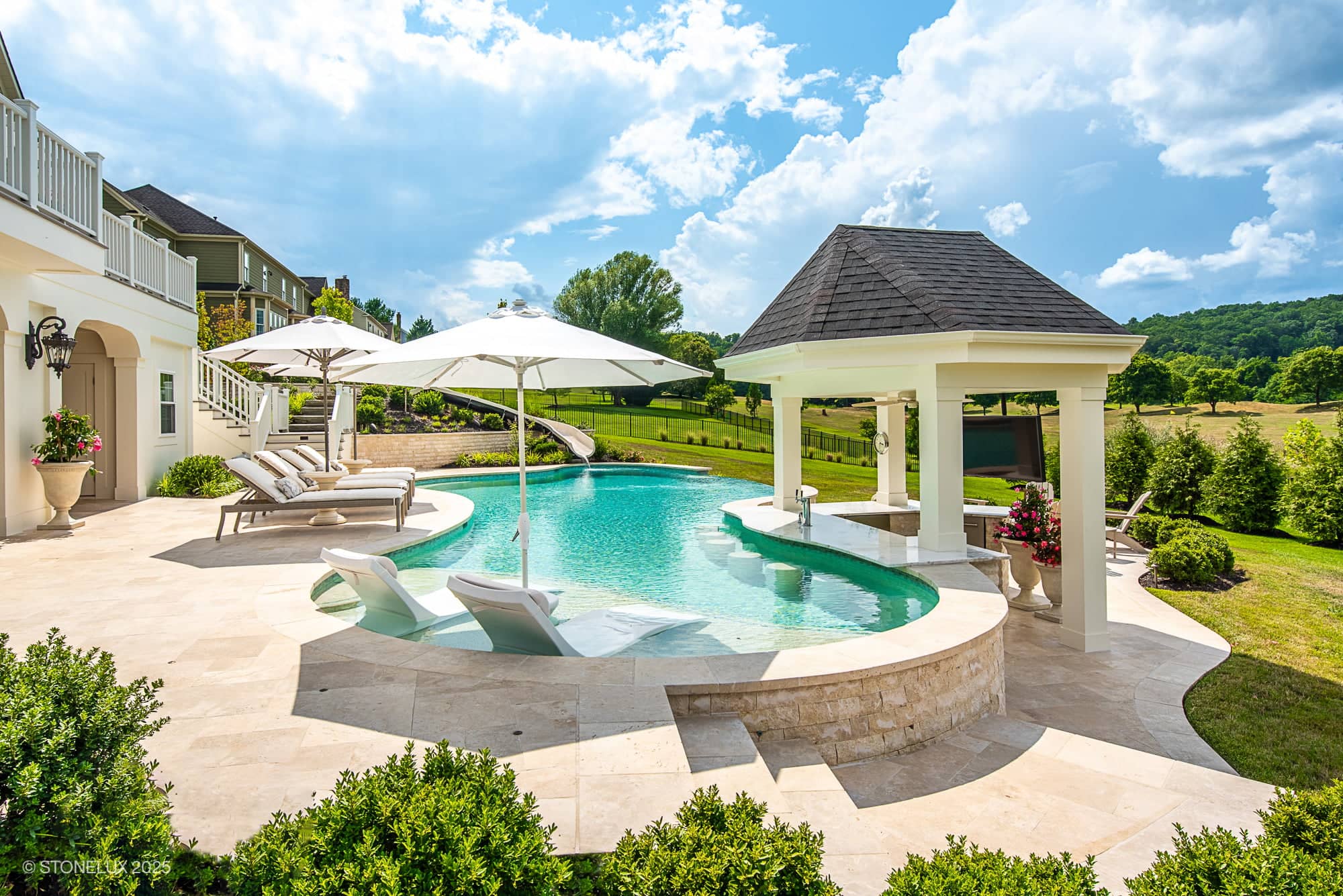 A luxurious backyard, the Leesburg Grecian Oasis – Ivory Travertine Pool Project, features a curved swimming pool with lounge chairs under white umbrellas, a small pavilion, lush landscaping, and views of grassy fields beneath a bright blue sky.