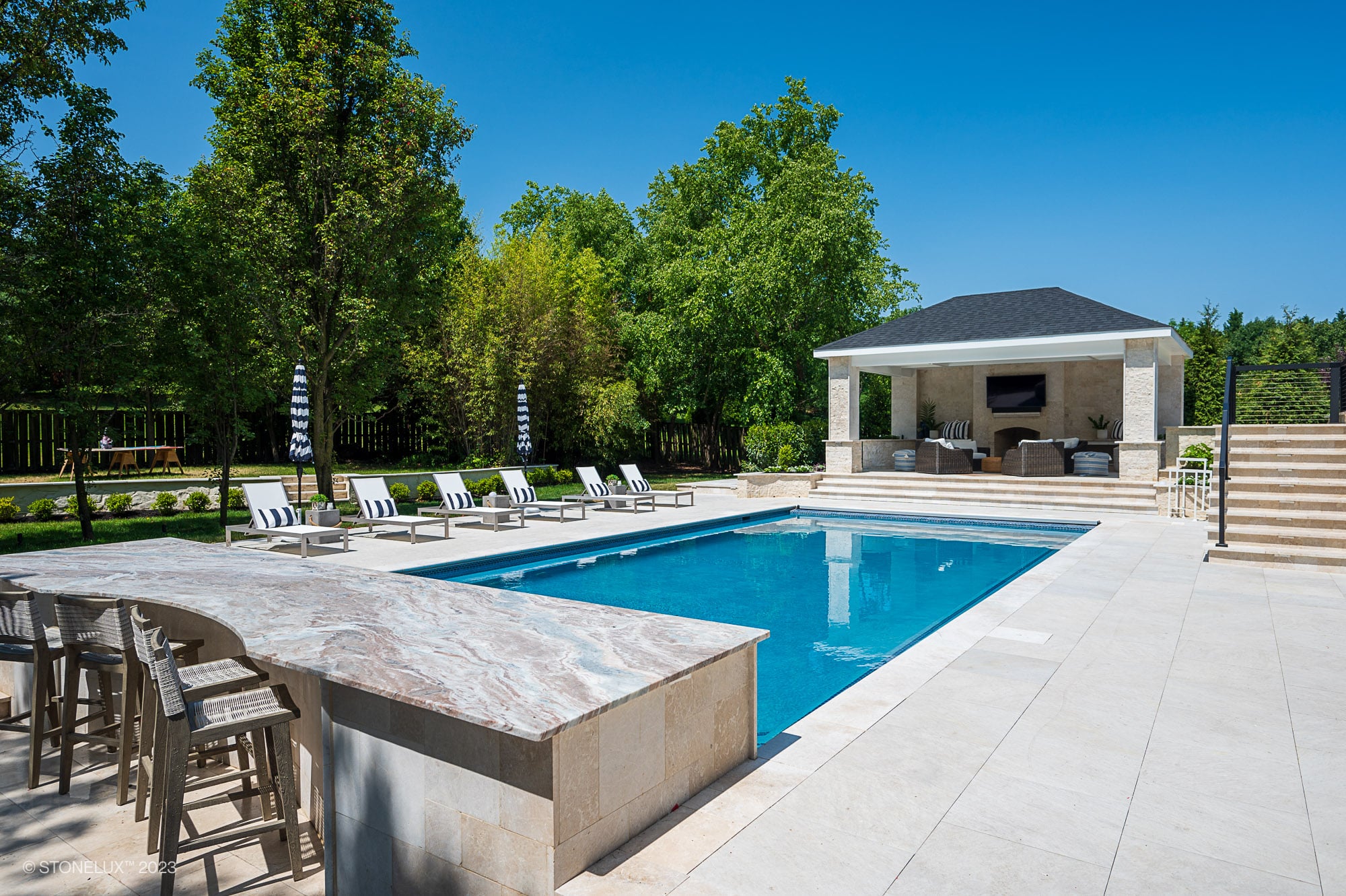 Luxury outdoor pool area with lounge chairs, a marble bar featuring White Harmony: Pera Cream Sandblasted Marble, stylish stools, and a covered pavilion with TV and seating—your porcelain poolside retreat surrounded by trees and a clear blue sky.