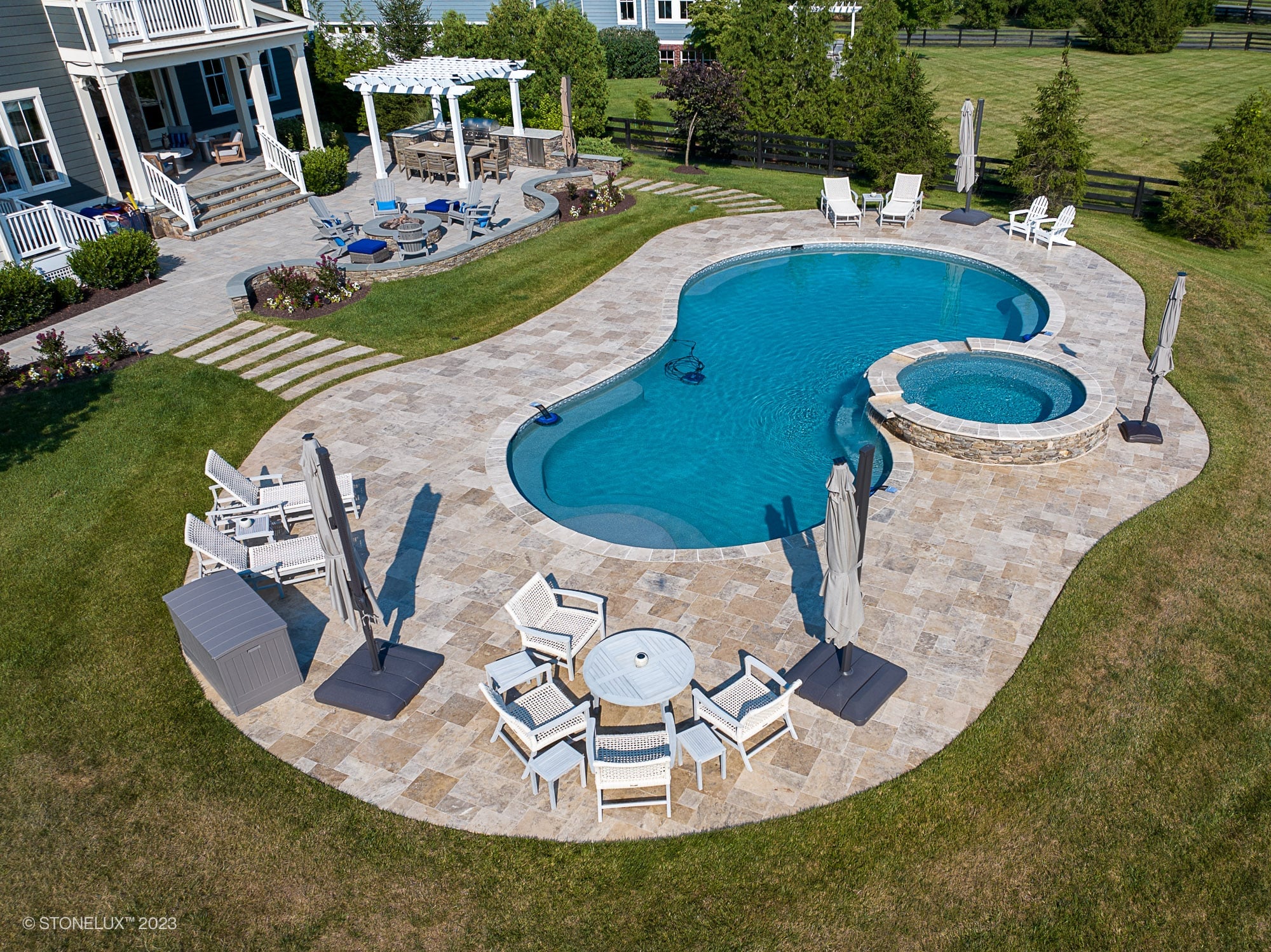 Aerial view of a backyard oasis featuring a Travertine Charm: Philly Travertine Pool with beige and blue accents, complemented by an attached hot tub, stone decking, lounge chairs, umbrellas, fire pit area, and lush landscaped grass.