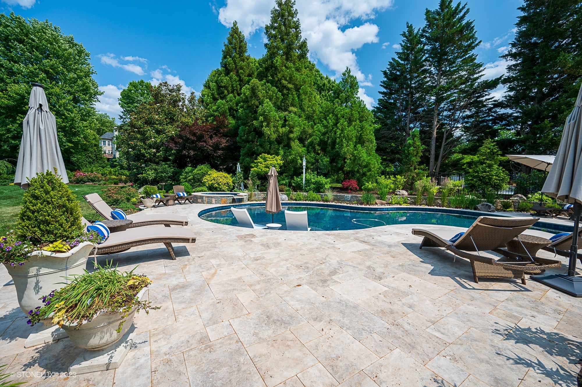 A backyard pool area with lounge chairs, umbrellas, and lush green landscaping on a sunny day. Travertine Serenity: a poolside palette of beige and silver light stone tiles enhances the patio amid trees and colorful plants.