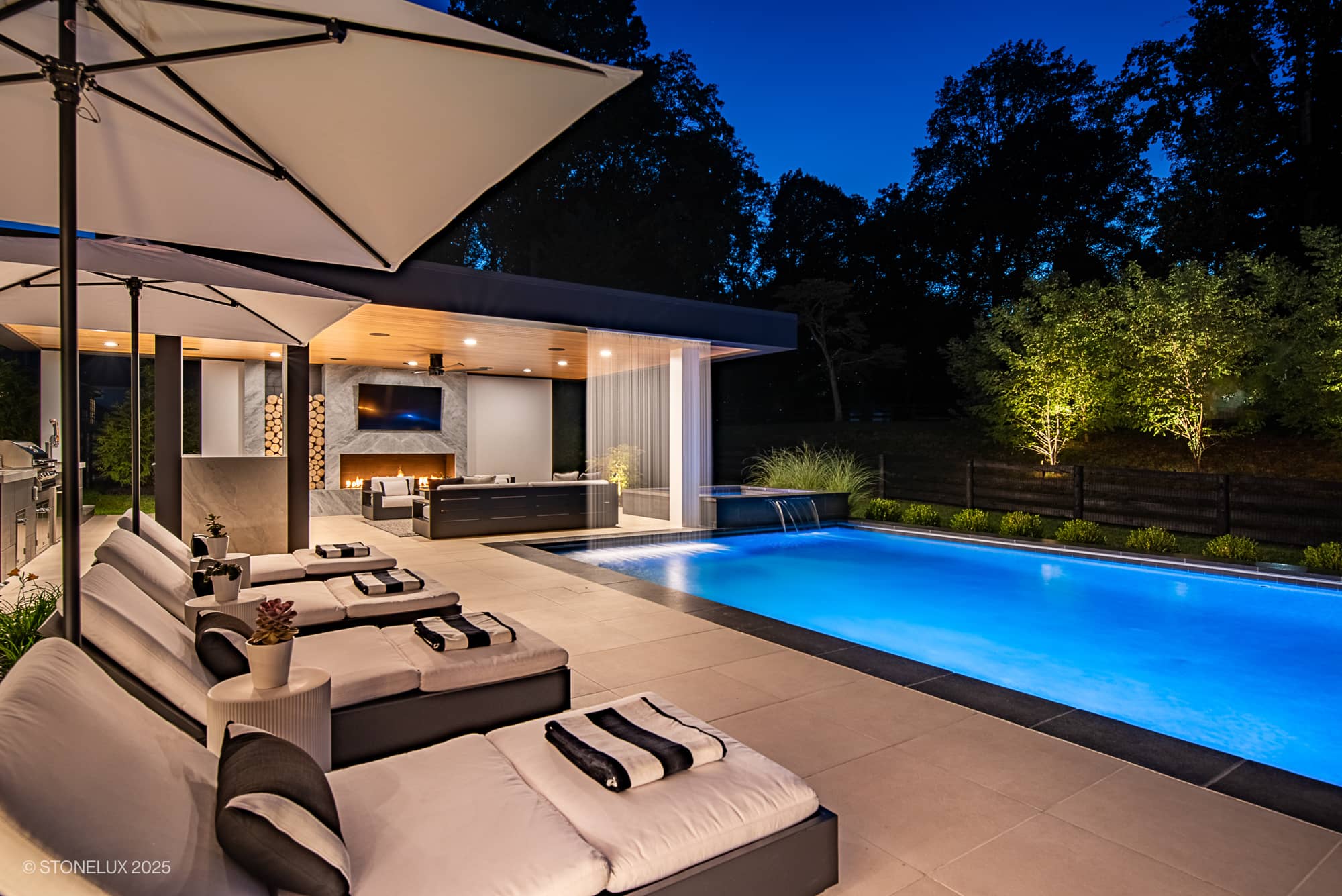 Experience the Aldie Modern Escape – Porcelain & Basalt Luxury Pool Pavilion: a stunning outdoor pool area at dusk, with an illuminated swimming pool, cushioned lounge chairs, large umbrellas, covered patio with TV, and landscaped trees.