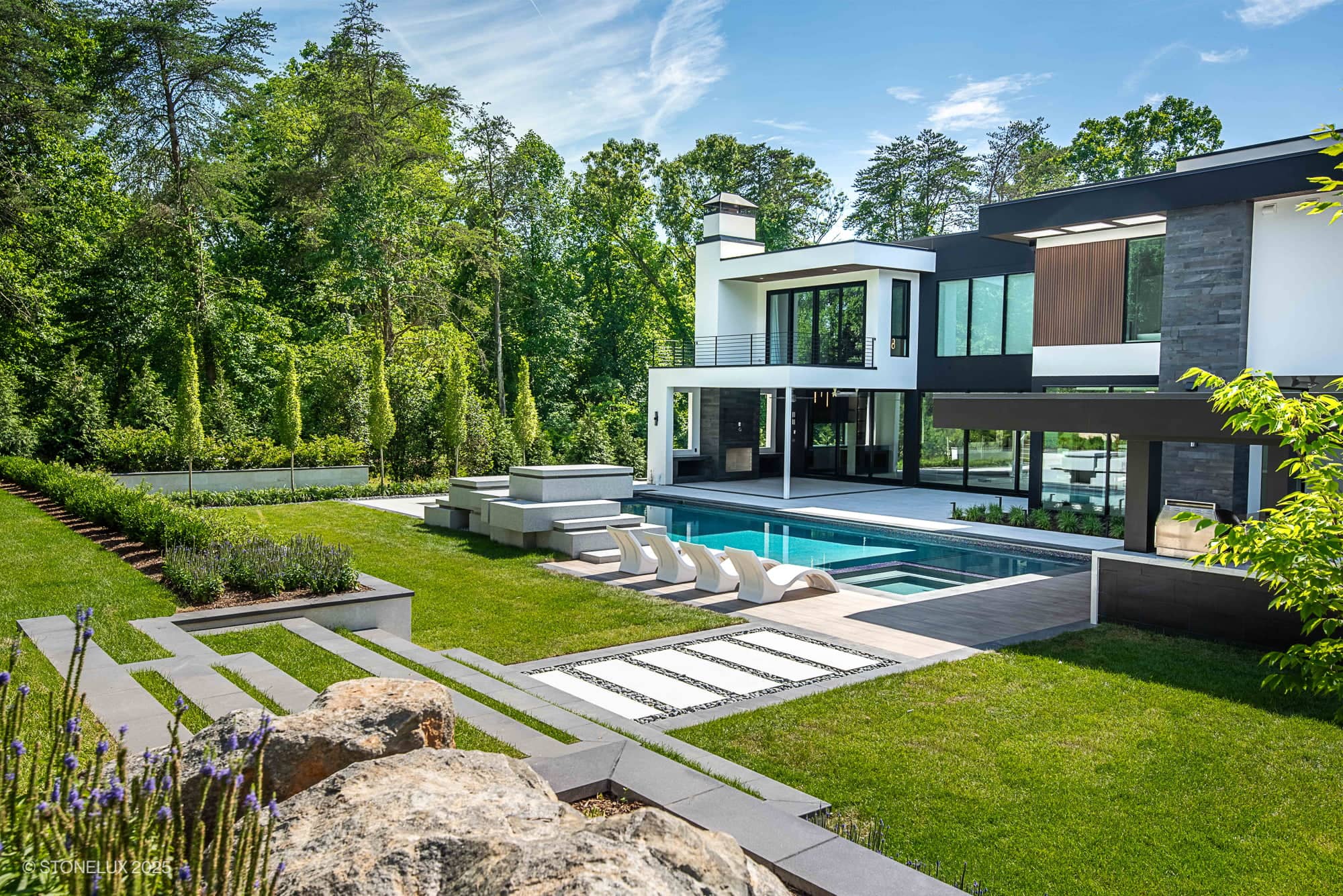 Modern Hillside Elegance – McLean Retreat featuring large windows, a pool, lounge chairs, hot tub, and manicured lawn with Overland Pearl Porcelain & Black Basalt accents, nestled among lush trees and landscaped gardens under a blue sky.