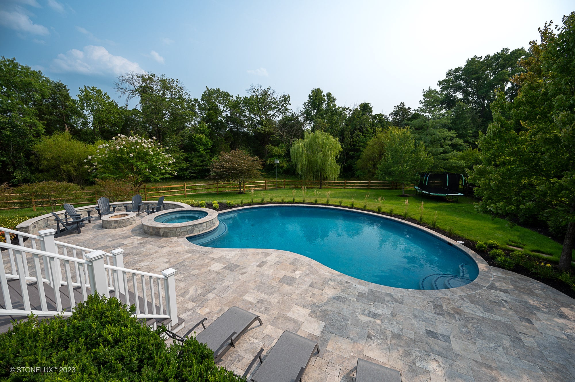 A backyard oasis featuring the Silver Splash: The Silver Travertine Pool Haven with an in-ground, kidney-shaped pool, hot tub, lounge chairs on a stone patio, white fenced staircase, lush lawn, trees, and a trampoline in the background.