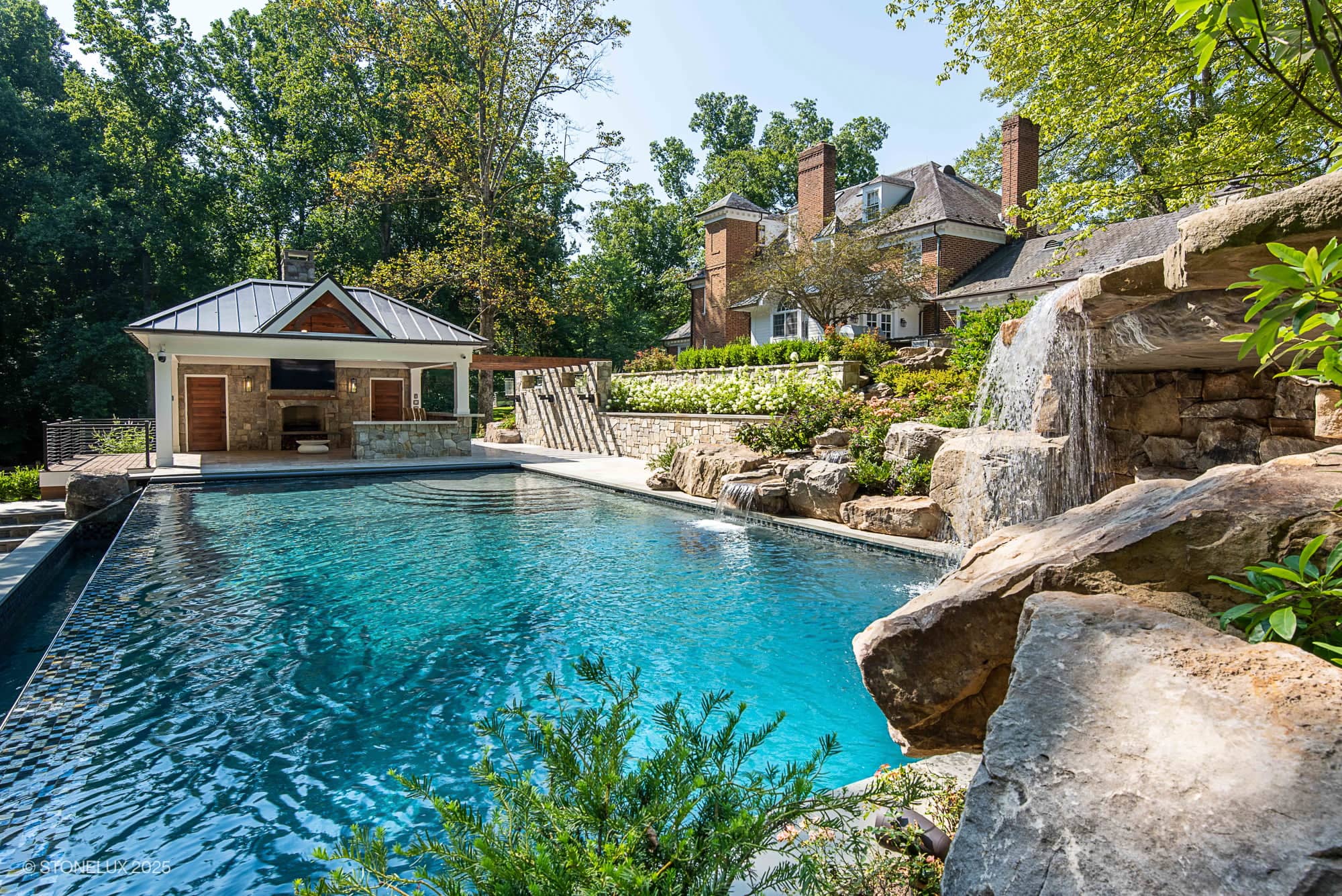 A luxurious backyard, dubbed the Clifton Cascade Retreat – Silver Travertine Oasis, features a large swimming pool with a waterfall surrounded by rocks and lush greenery. A pool house with a TV and lounge area sits in the background near a brick home.