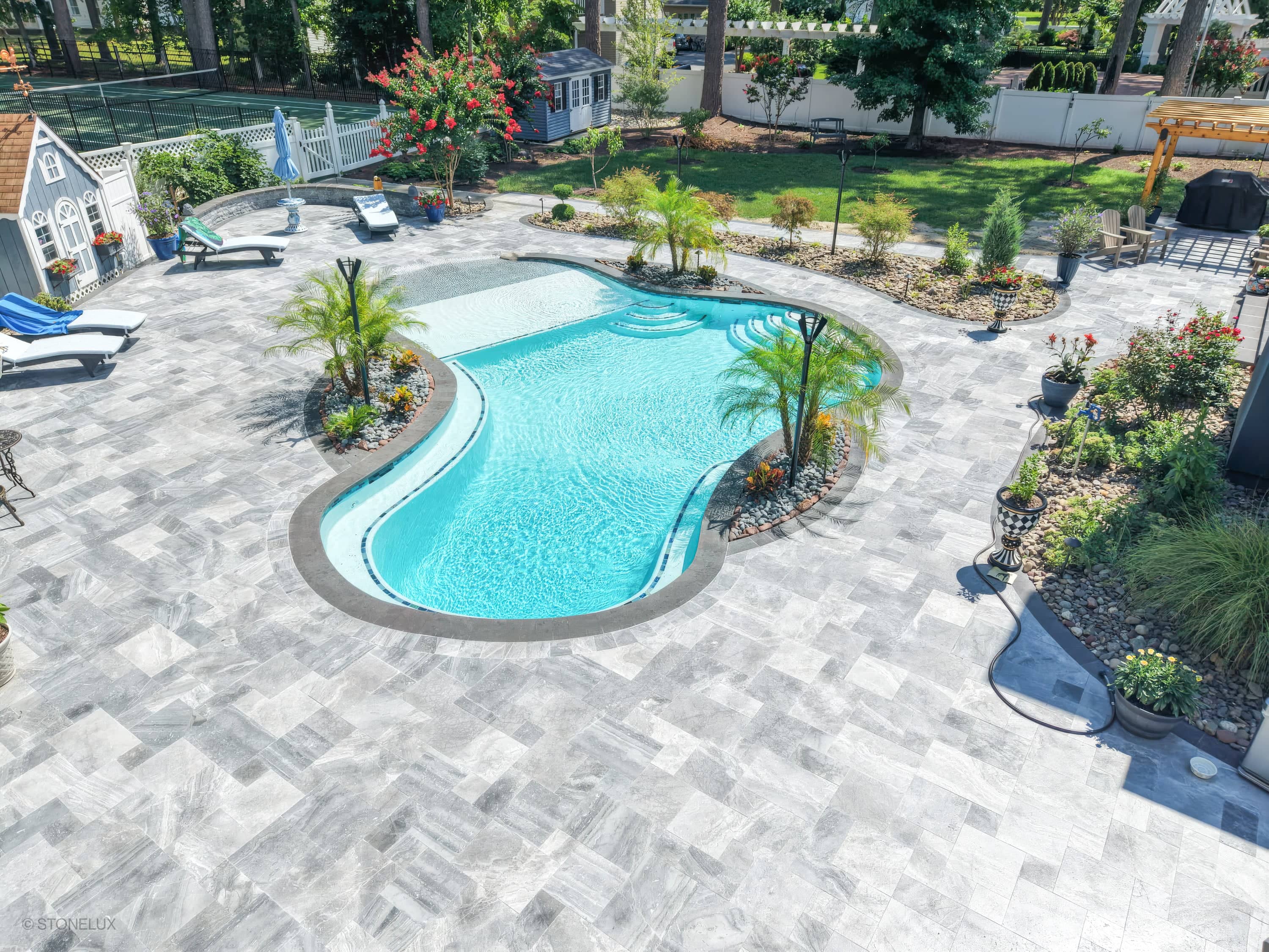 Aegean Grey Marble Pool Retreat in Salisbury, MD features a sleek pool with black basalt coping. The blue water contrasts beautifully with the grey marble pavers. Complementary tropical landscaping and elegant patio furniture enhance the modern outdoor...