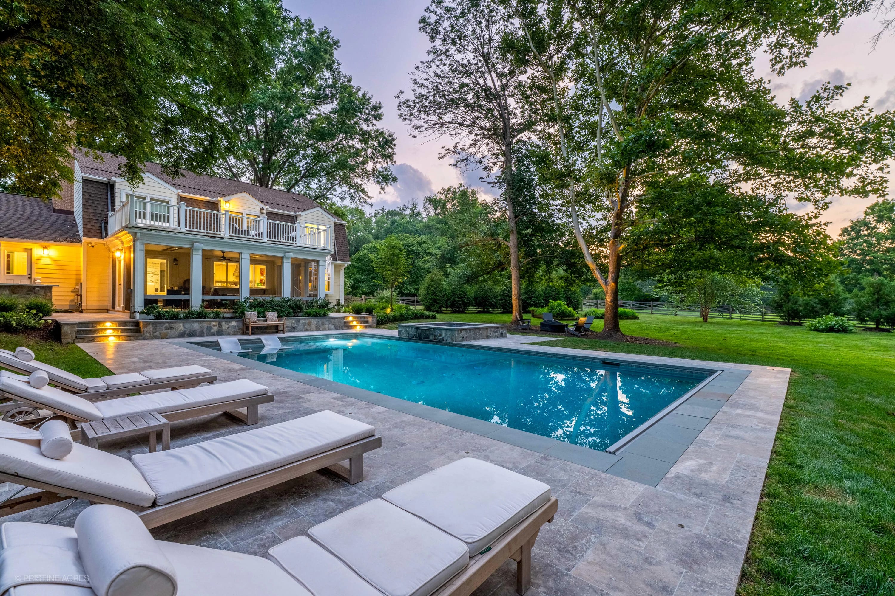 Elegant Vienna Pool Retreat featuring Silver Travertine collection pavers and Bluestone coping. The modern pool reflects blue water, surrounded by white lounge chairs and lush greenery. Warm lighting enhances the tranquil backyard setting.