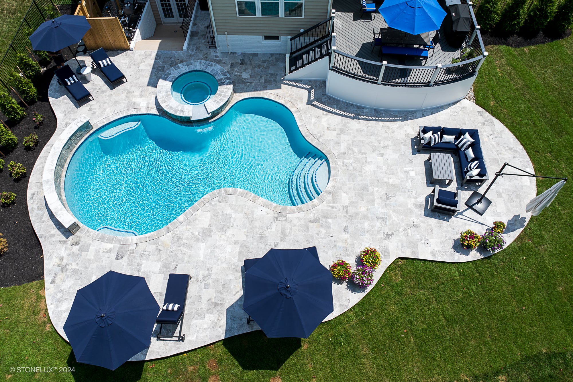 Aerial view of a backyard pool and patio project in Ashburn, VA, featuring a curvy swimming pool with attached hot tub, lounge chairs, umbrellas, grassy lawn, and elegant Tundra Grey Marble accents beside the stone patio.