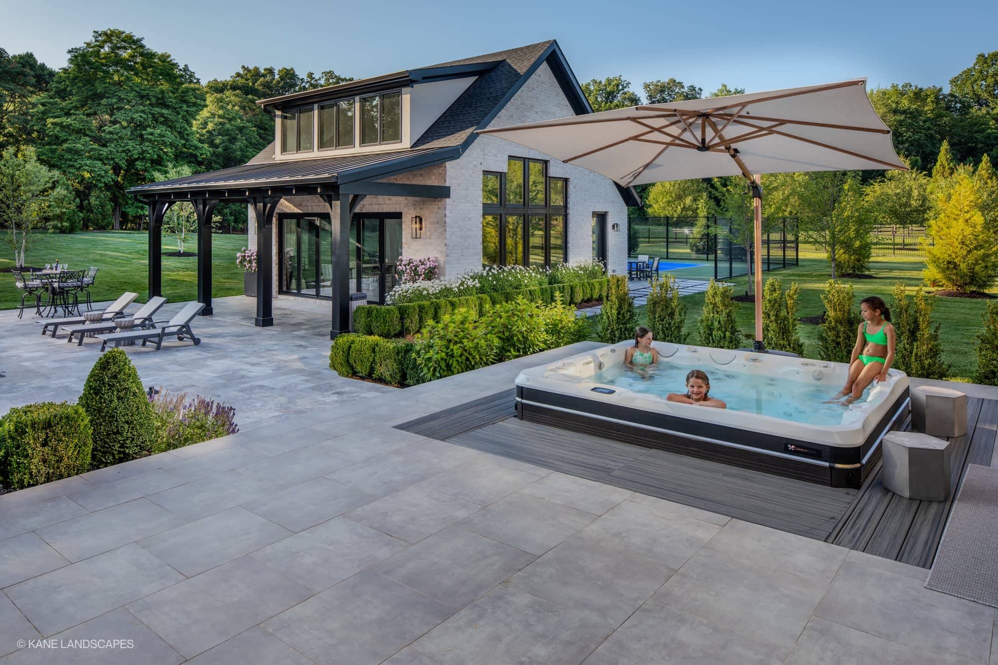Modern Oasis at Grenata Preserve in Leesburg, VA features Silver Travertine Premium pavers. A sleek patio with lush green landscaping surrounds a spa. Modern architecture complements the serene setting, with grey pavers enhancing the stylish outdoor fu...
