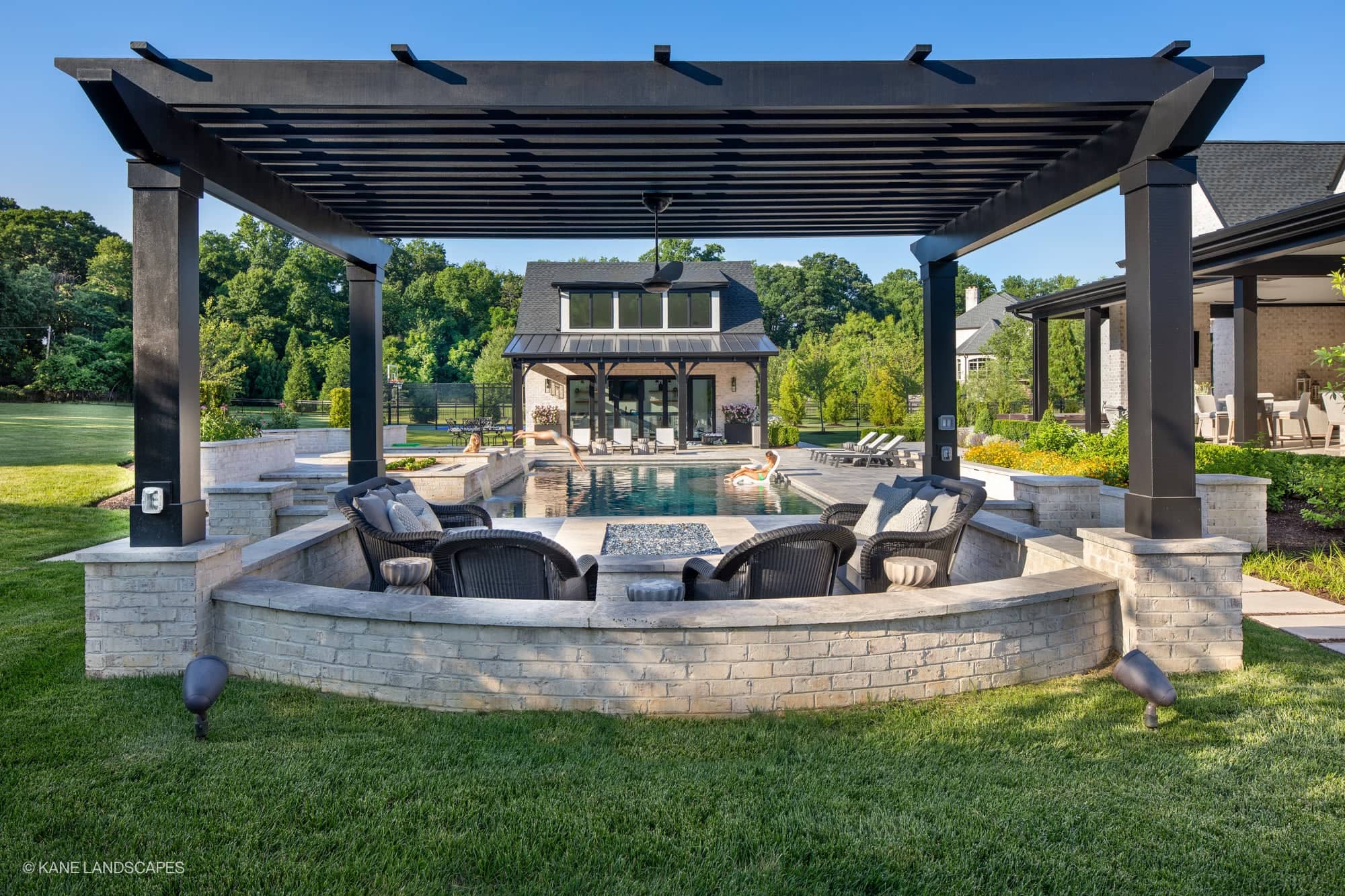 Modern Oasis at Grenata Preserve features Silver Travertine Premium pavers in a contemporary Leesburg, VA backyard. The sleek pool reflects the blue sky, surrounded by lush greenery and stylish black pergola. Cozy wicker seating complements the elegant...
