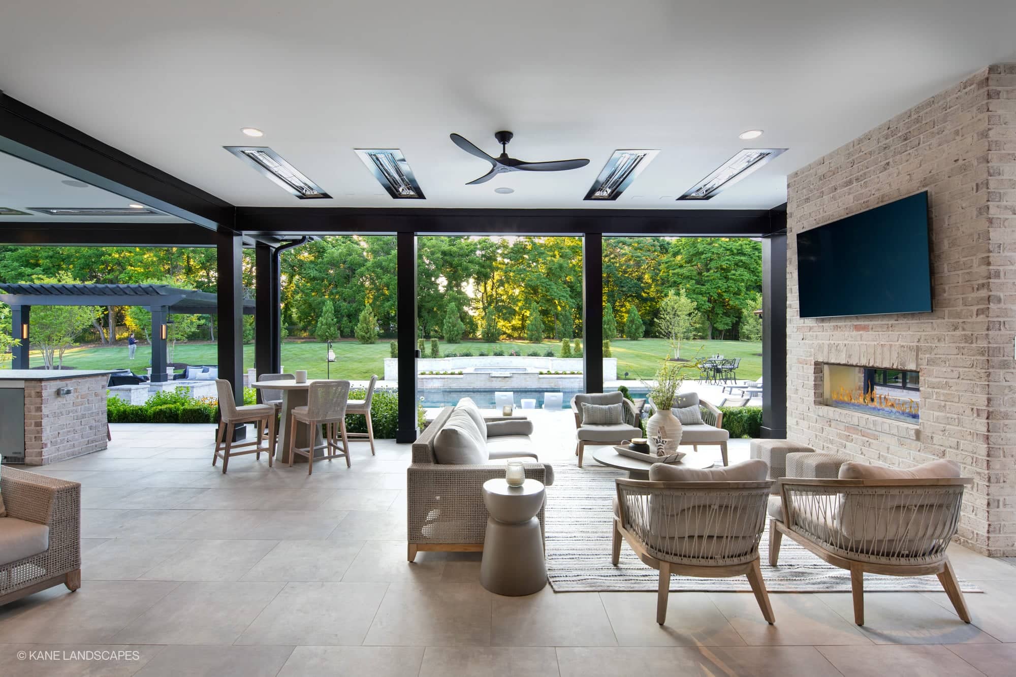 Modern Oasis at Grenata Preserve in Leesburg, VA features Silver Travertine Premium pavers. The sleek patio connects to a lush garden with a pool. Stylish outdoor furniture and a cozy fireplace enhance this modern hardscape project.