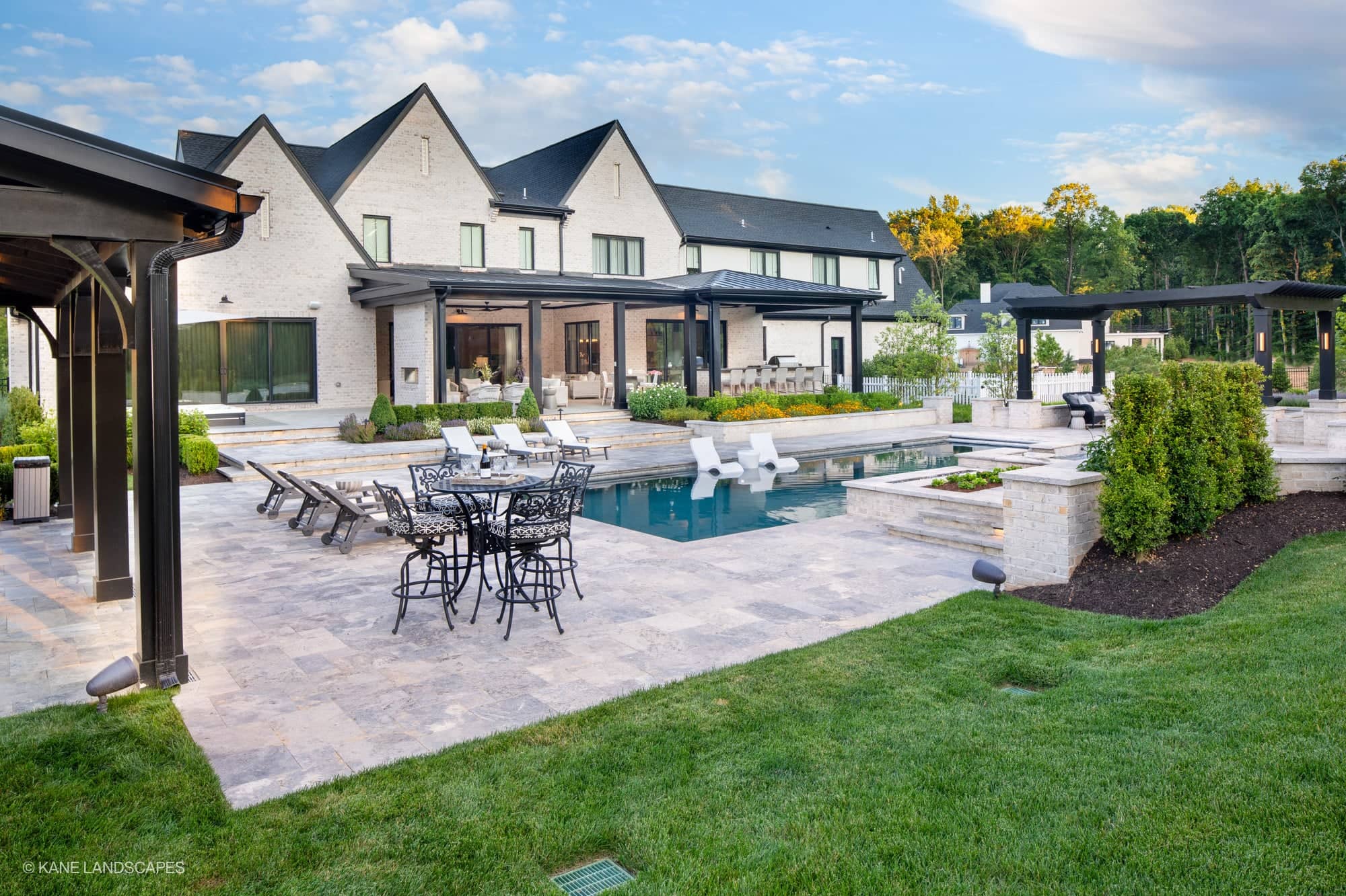 Modern Oasis at Grenata Preserve in Leesburg, VA showcases Silver Travertine Premium from STONELUX. The modern design features elegant travertine pavers around a serene blue pool, complemented by white lounge chairs, black metal outdoor furniture, and ...