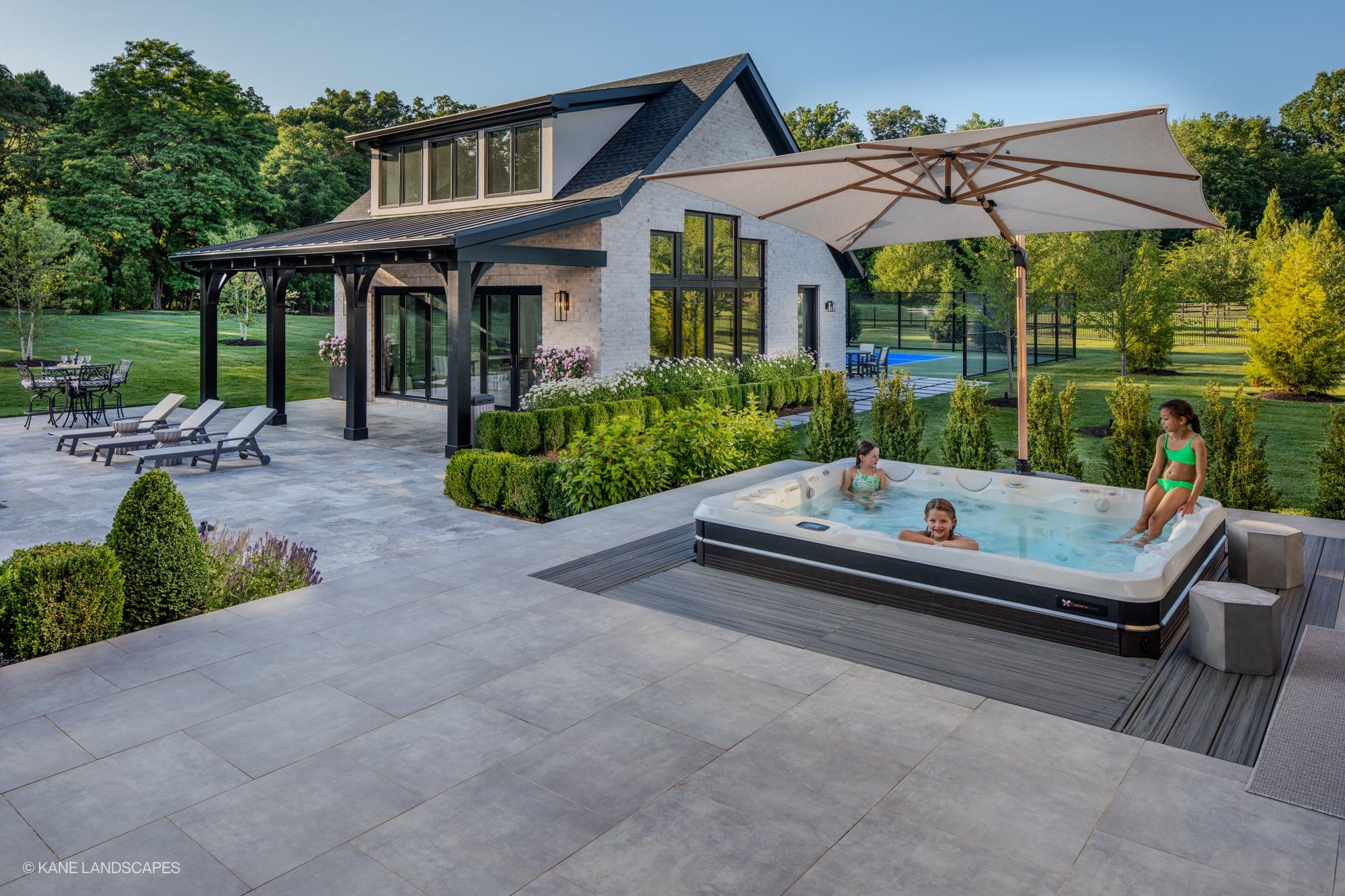 Modern Oasis at Grenata Preserve in Leesburg, VA features Silver Travertine Premium pavers. A sleek patio with lush green landscaping surrounds a spa. Modern architecture complements the serene setting, with grey pavers enhancing the stylish outdoor fu...