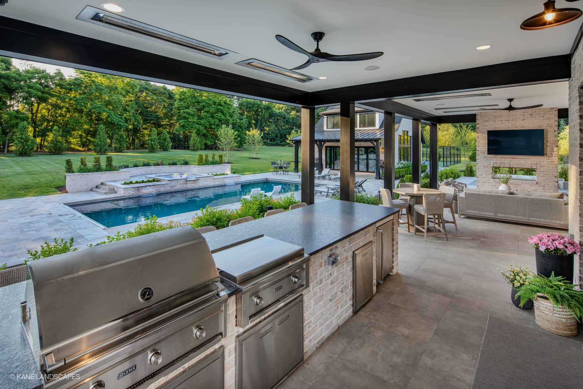 Modern Oasis at Grenata Preserve in Leesburg, VA, featuring Silver Travertine Premium pavers. A sleek pool with grey coping complements the clear blue water. Surrounded by lush greenery, a contemporary patio with outdoor kitchen, seating, and elegant l...