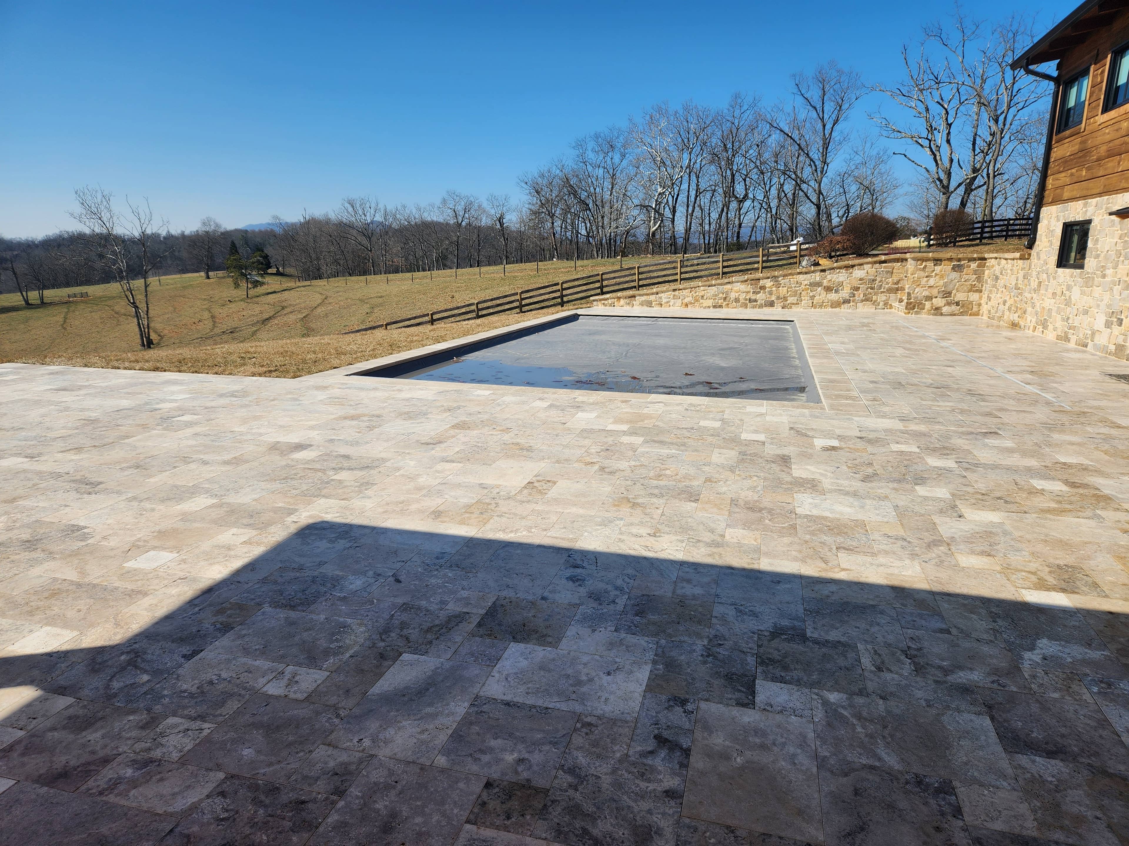 A large stone patio by a wooden and stone building showcases a covered rectangular pool, part of the VA Pool & Patio Project – Philly Travertine Elegance in Winchester. A fence borders open grassy fields with leafless trees under a clear blue sky.