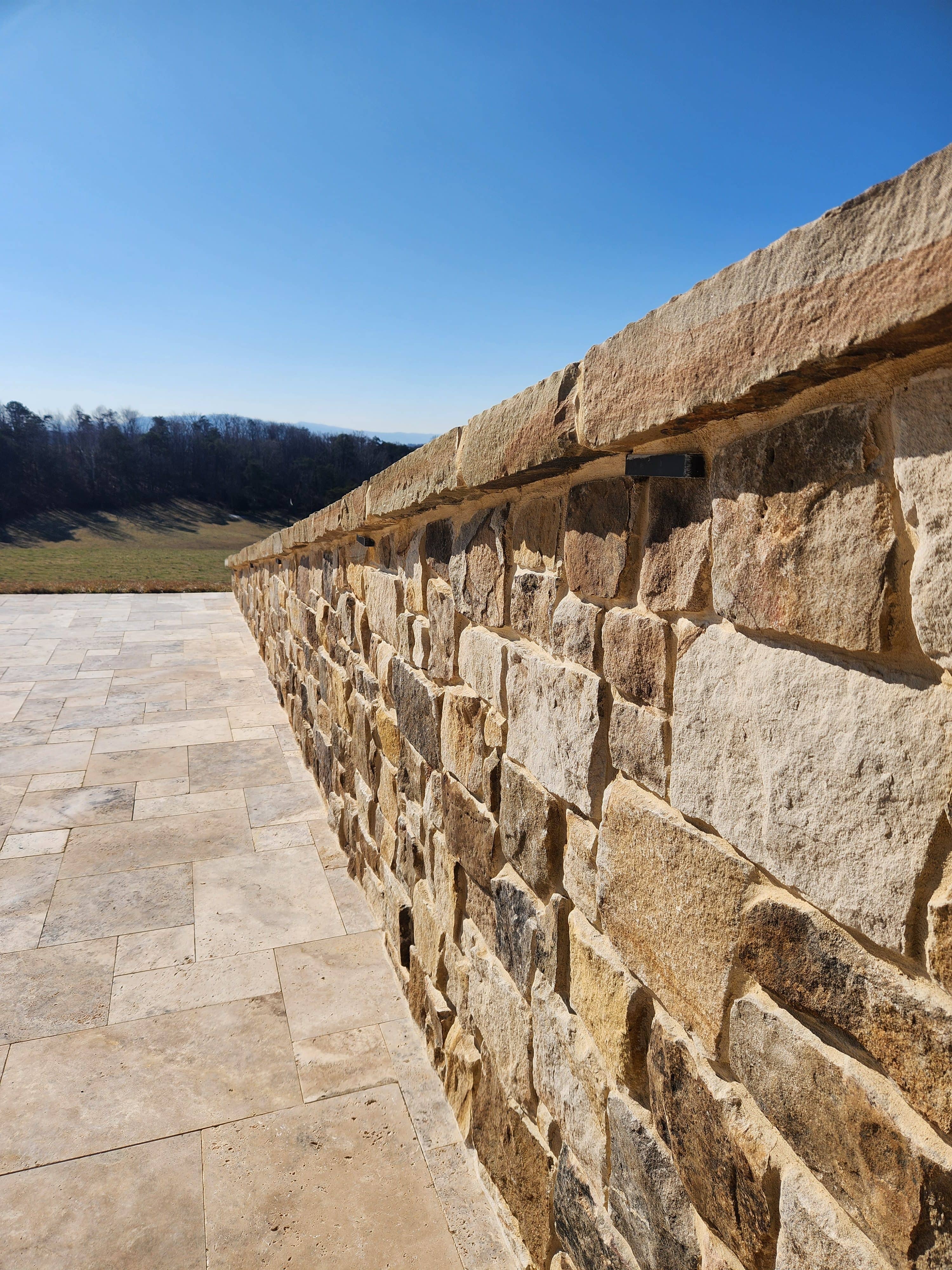 A close-up view of a stone wall and stone-paved ground showcases VA Pool & Patio Project – Philly Travertine Elegance under a clear blue sky, with grassy fields and distant trees in the Winchester landscape.