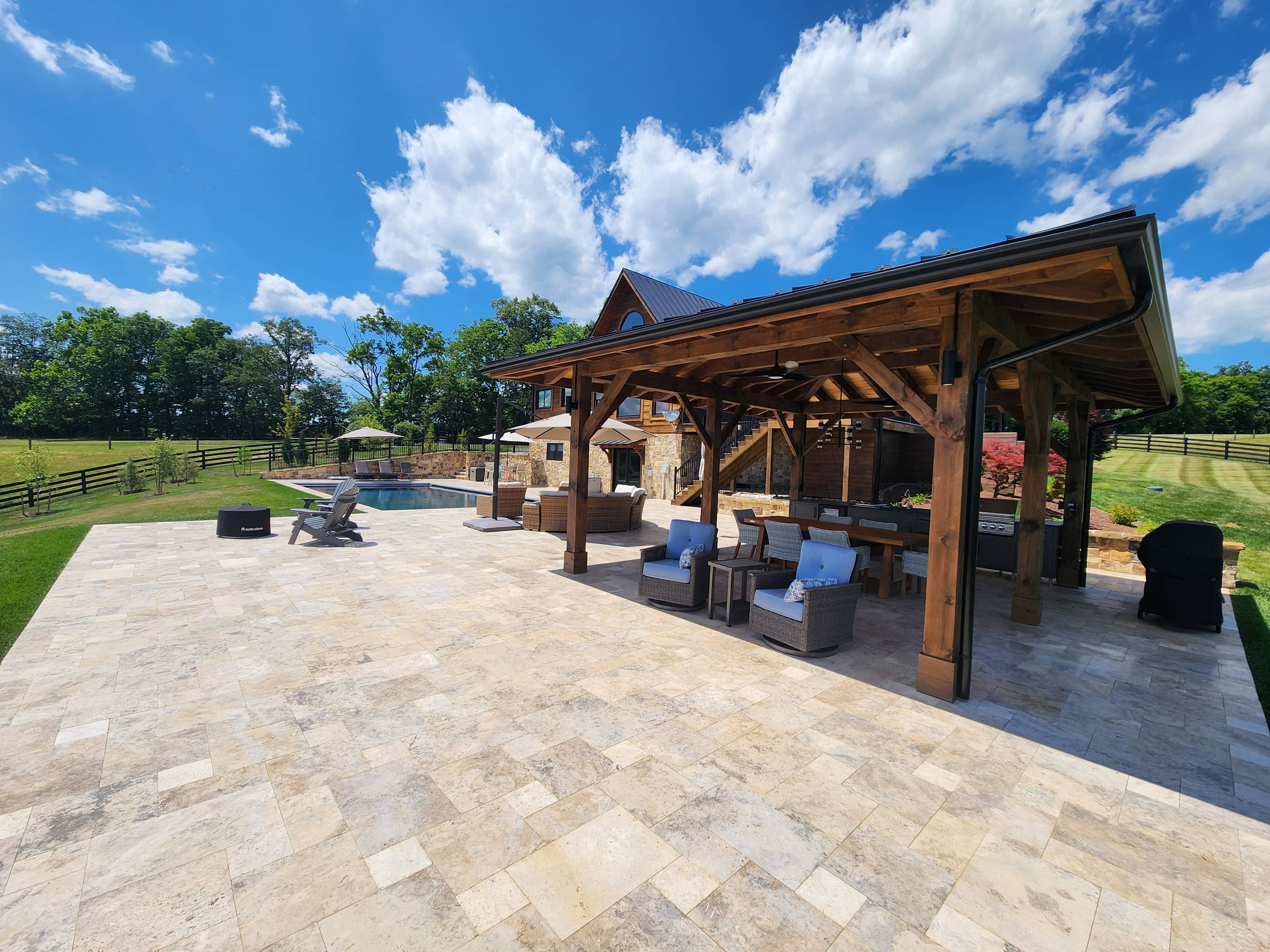 Spacious outdoor patio with stone tiles and a wooden pergola, featuring seating areas, grill, and pool—part of the VA Pool & Patio Project – Philly Travertine Elegance. Surrounded by green lawn, trees, and a wooden fence under a bright blue sky.