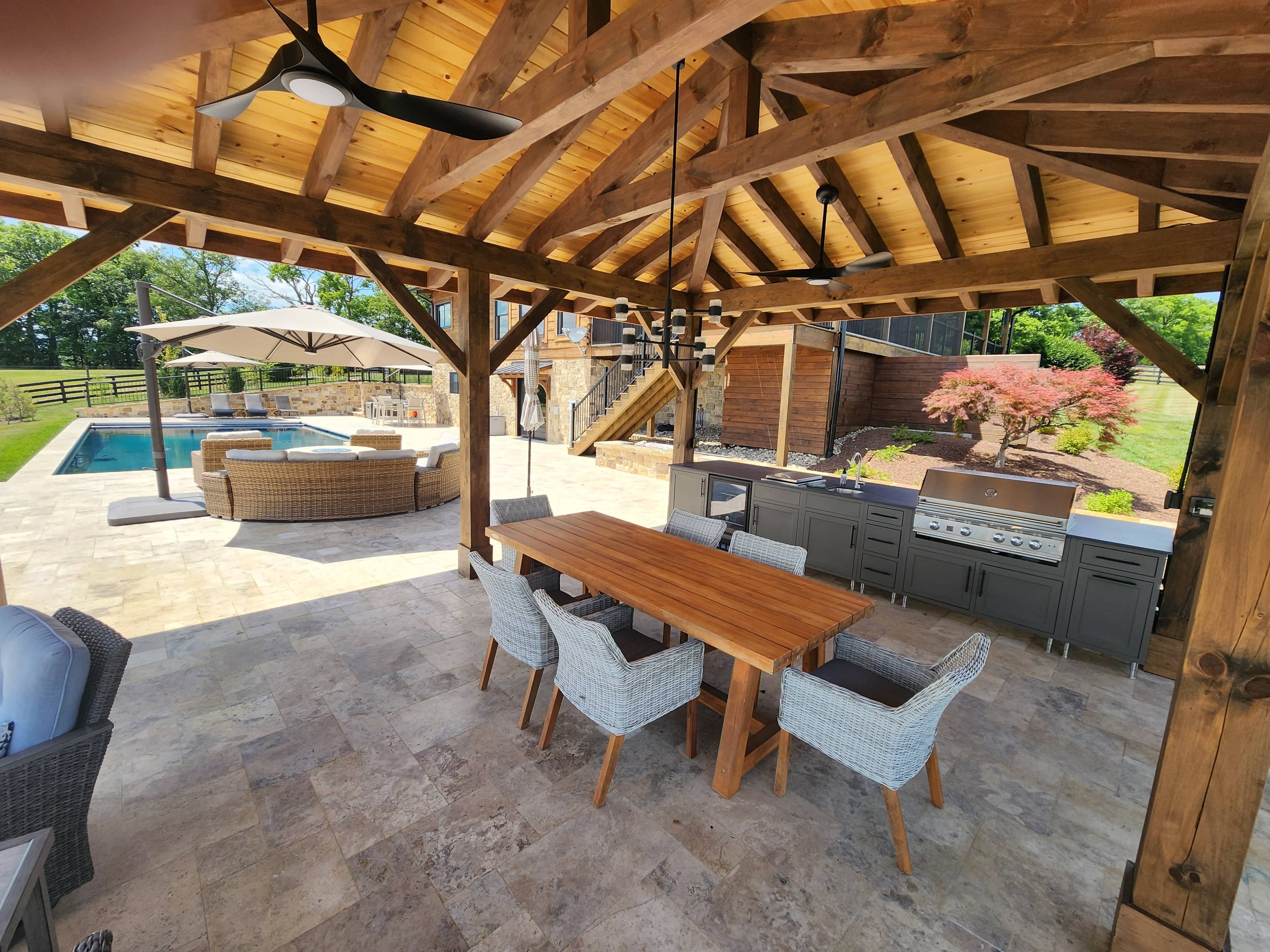 A covered outdoor patio at the VA Pool & Patio Project – Philly Travertine Elegance in Winchester features a wooden dining table with wicker chairs, built-in grill and kitchen, ceiling fan, and overlooks a pool area surrounded by lush greenery.