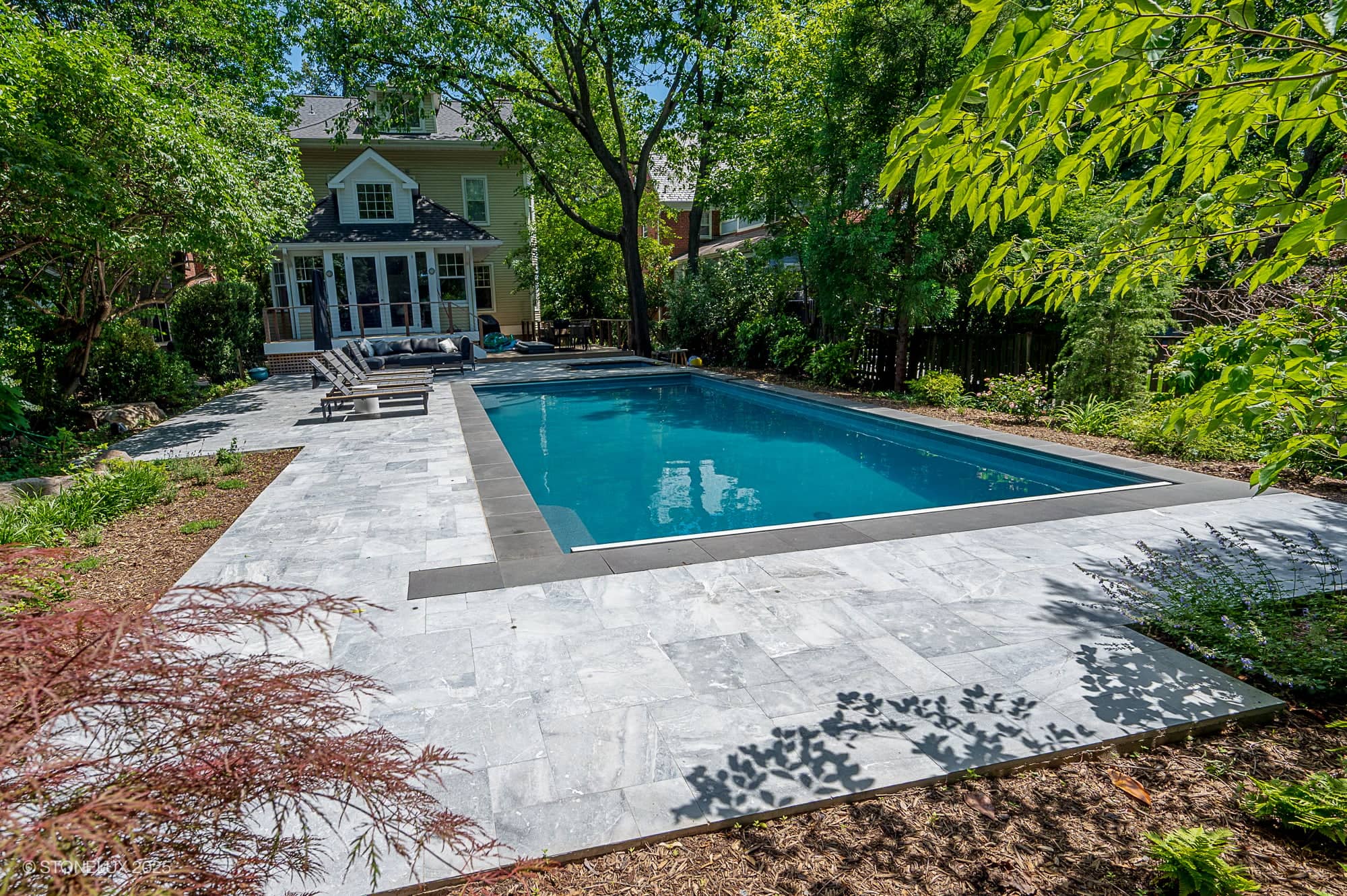 Experience urban serenity in this backyard oasis featuring the Urban Serenity Pool Project—an elegant rectangular pool edged with Aegean Grey Marble & Black Basalt, surrounded by a gray stone patio, lounge chairs, and lush greenery.