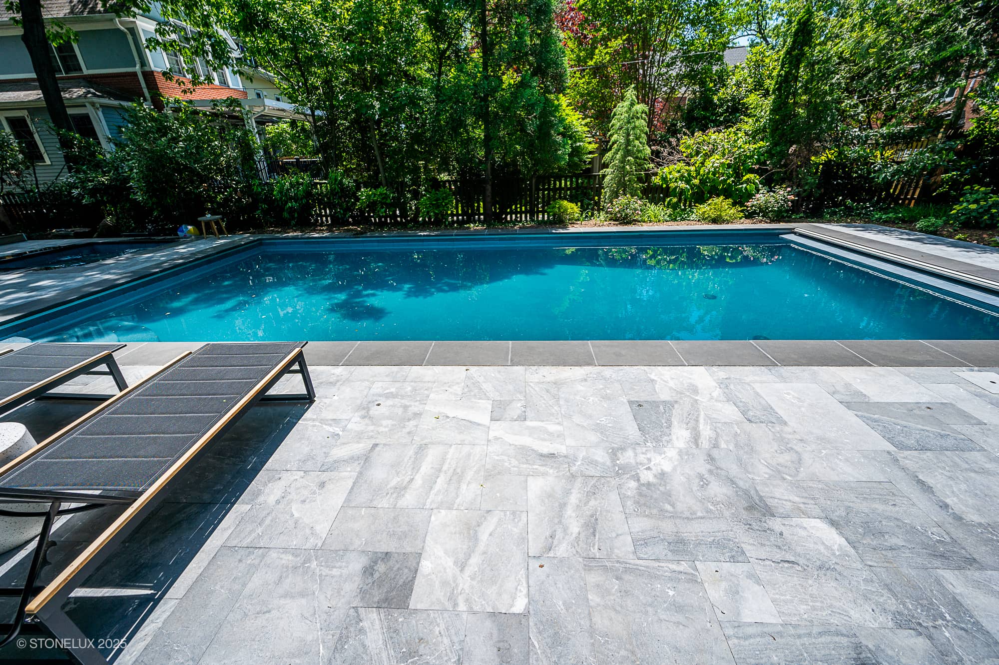 A rectangular backyard swimming pool with clear blue water, part of the Urban Serenity Pool Project, features Aegean Grey Marble and Black Basalt tiles, two lounge chairs on the left, and lush green trees and plants in the background.