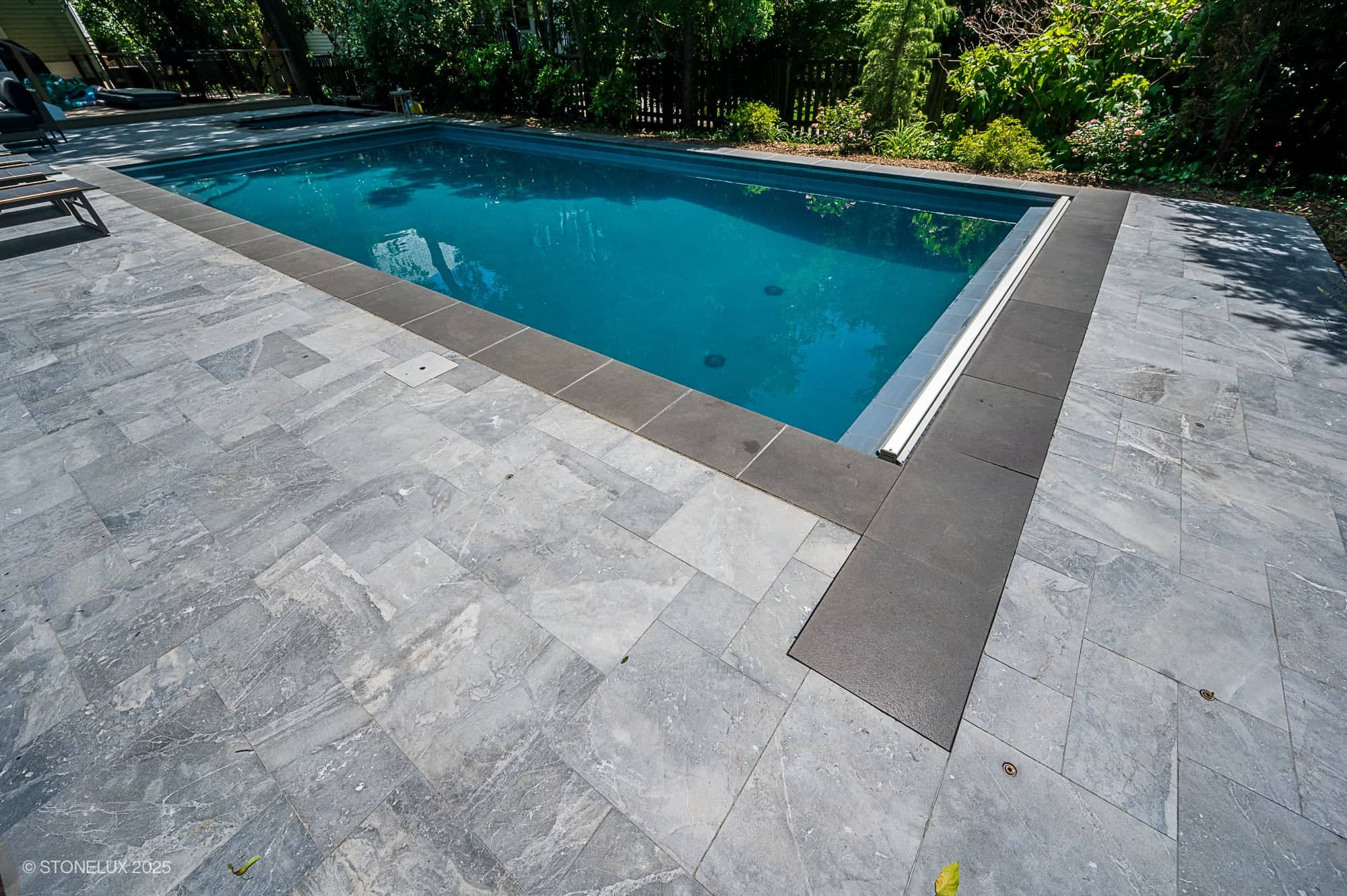 A modern backyard swimming pool, part of the Urban Serenity Pool Project, features clear blue water, Aegean Grey Marble tiles with a Black Basalt border, and is set amid lush green plants and trees for a tranquil retreat.