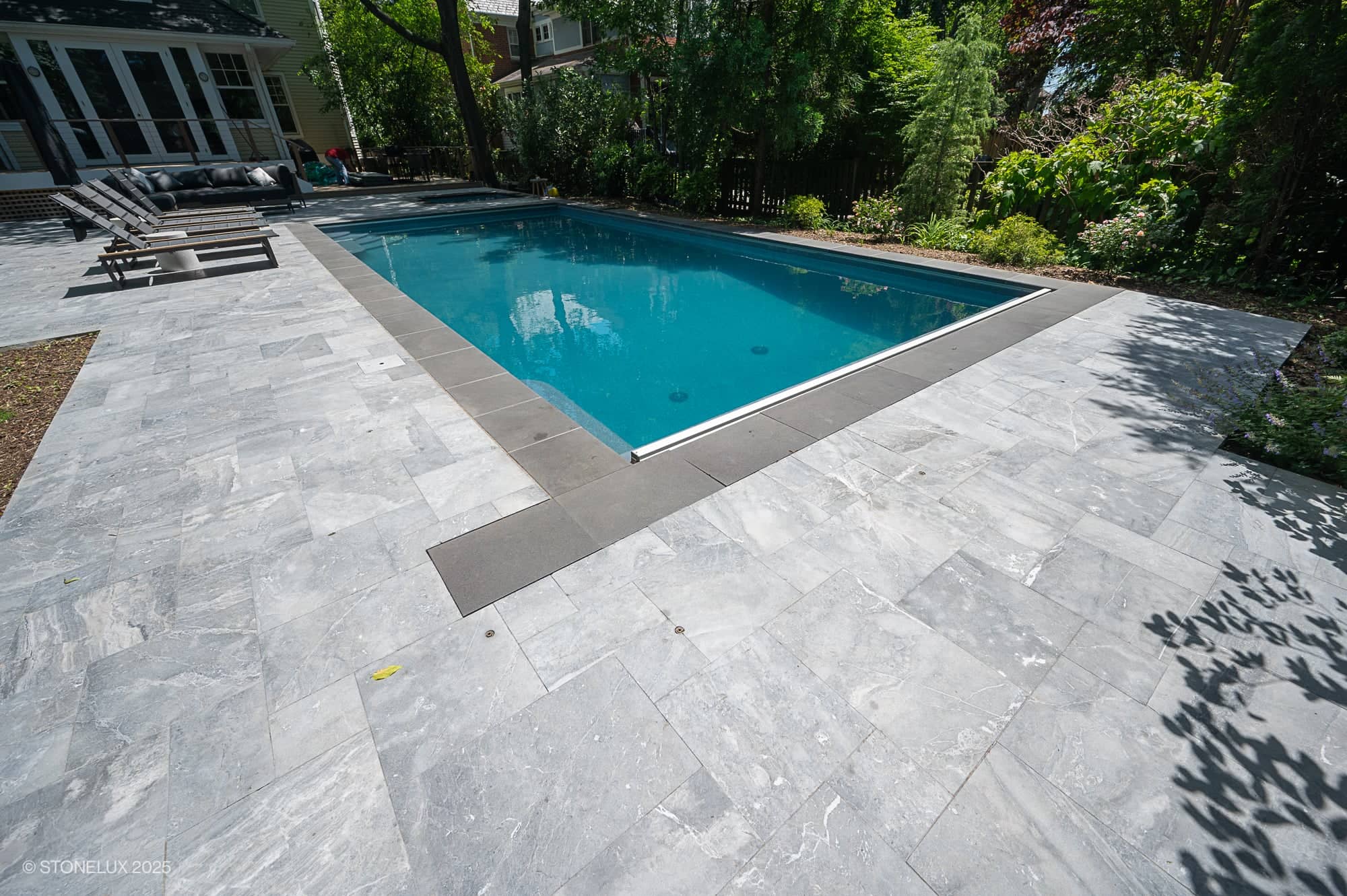 A rectangular outdoor swimming pool, part of the Urban Serenity Pool Project, features clear blue water and an Aegean Grey Marble & Black Basalt patio. Lounge chairs sit to the left, with trees and greenery in the sunny background.