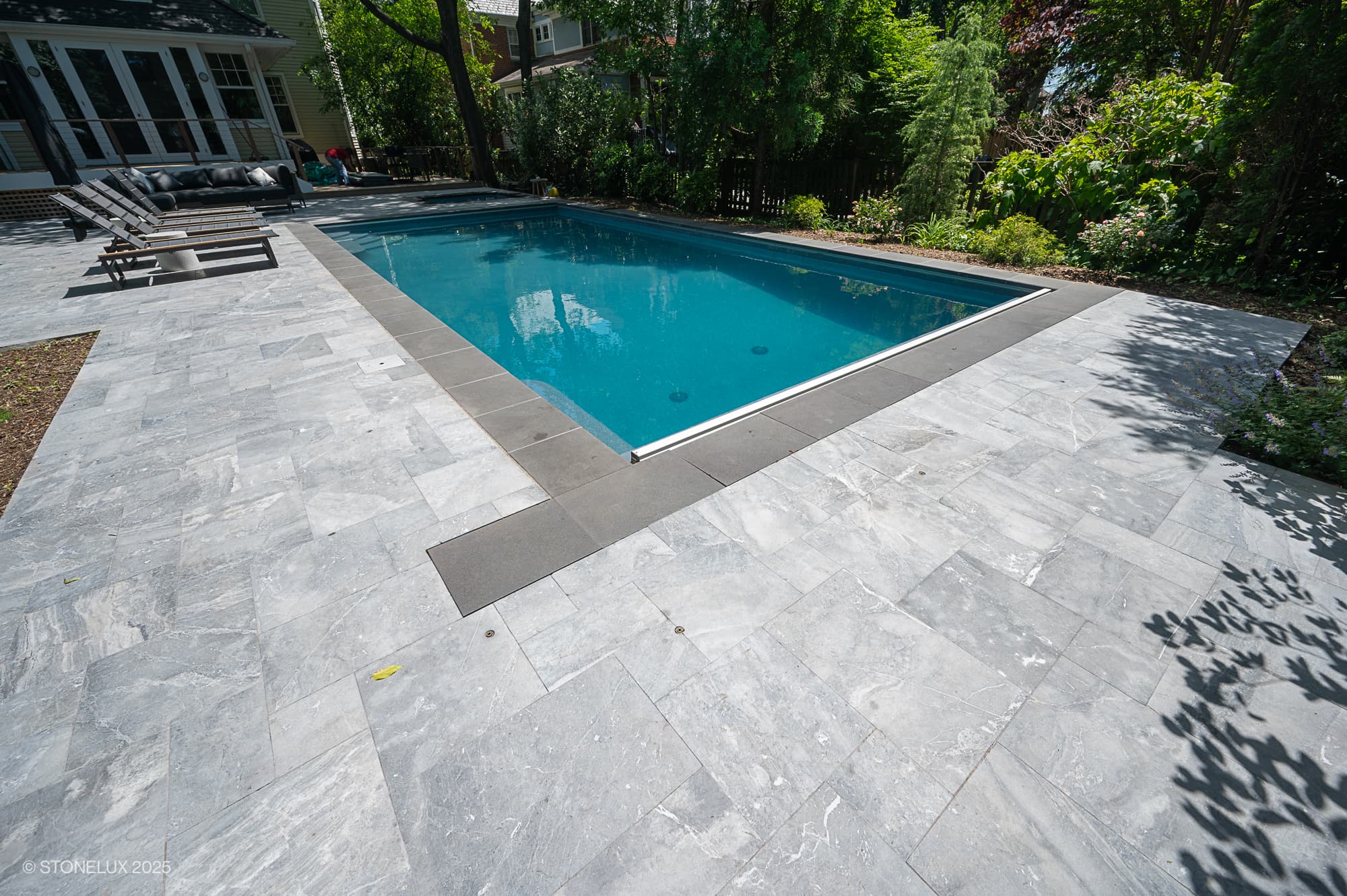 A rectangular outdoor swimming pool, part of the Urban Serenity Pool Project, features clear blue water and an Aegean Grey Marble & Black Basalt patio. Lounge chairs sit to the left, with trees and greenery in the sunny background.