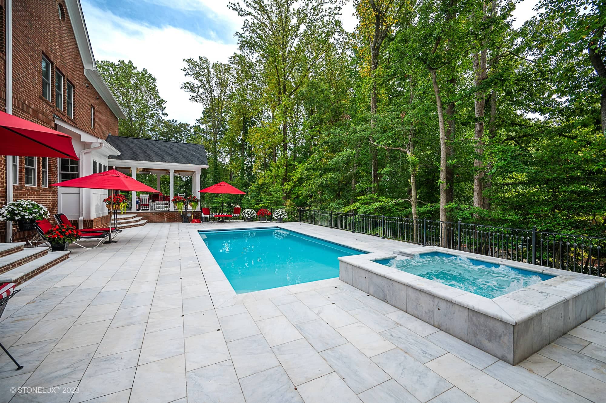 A backyard with a rectangular swimming pool and adjacent hot tub, surrounded by a light stone patio in White Whisper: The Pristine Afyon Marble Pool Elegance. Red umbrellas and lounge chairs sit near a brick house, framed by dense green trees.